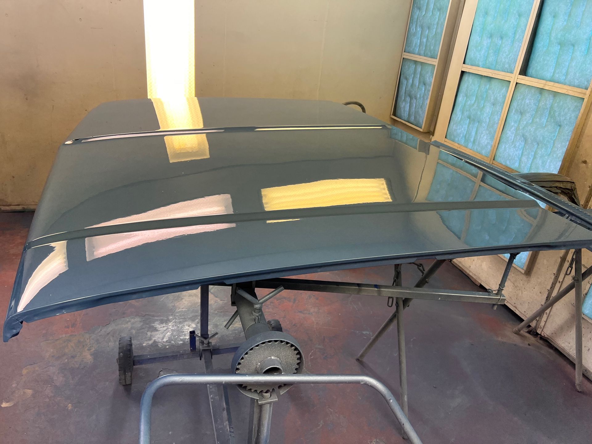 Dark gray car hood on a stand in a workshop, with a roll-up door and window reflections nearby.