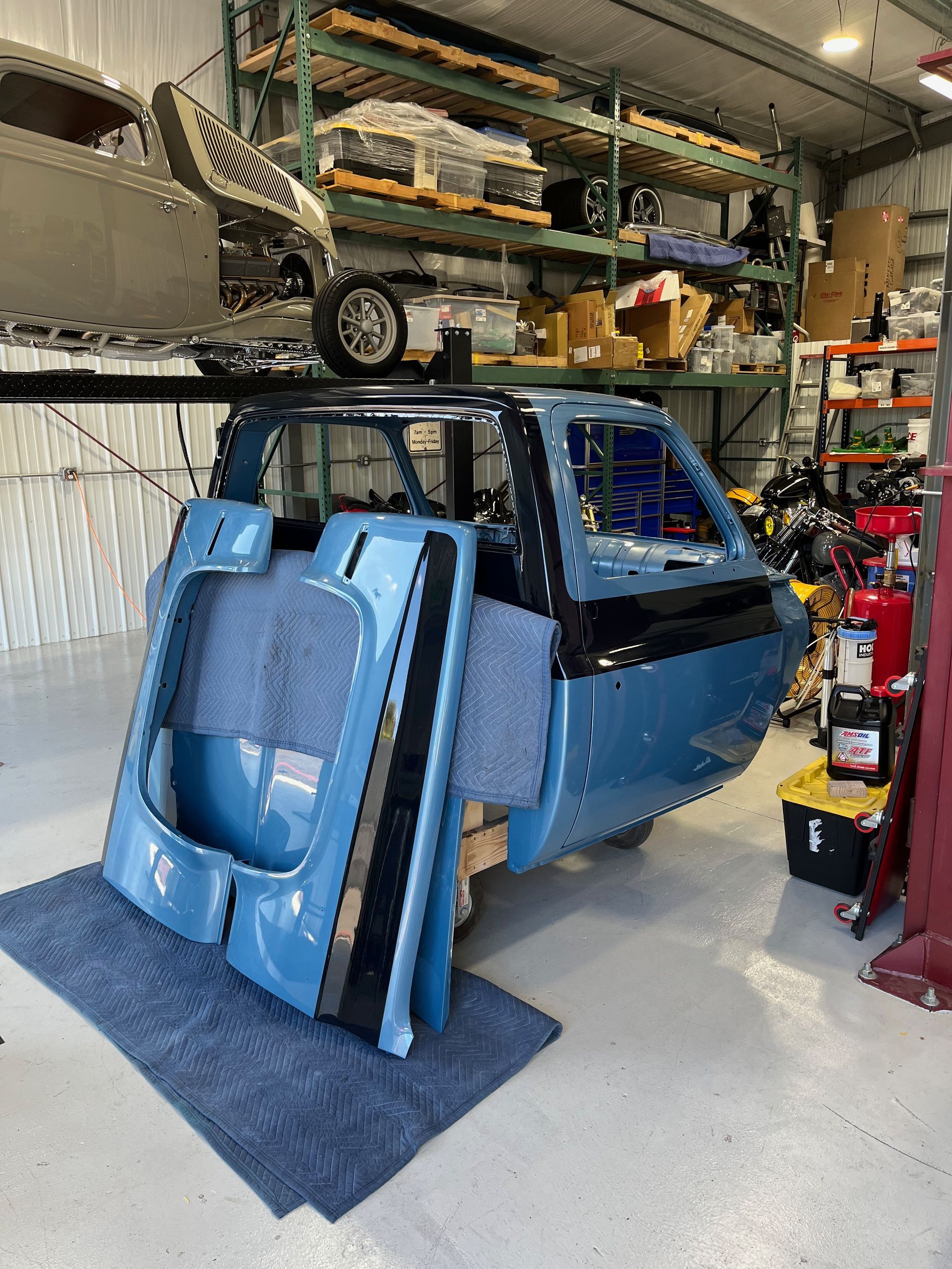 Blue van body with doors removed in a garage workshop, surrounded by tools and vehicle parts.