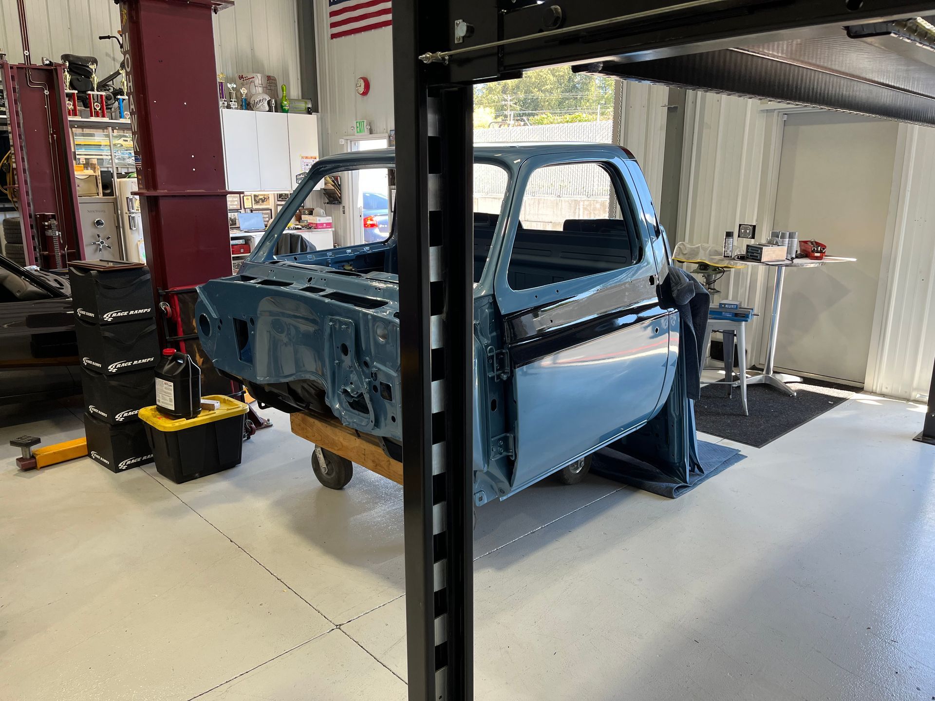 Blue pickup truck body in an auto repair shop on a lift, with red machinery and garage equipment around it