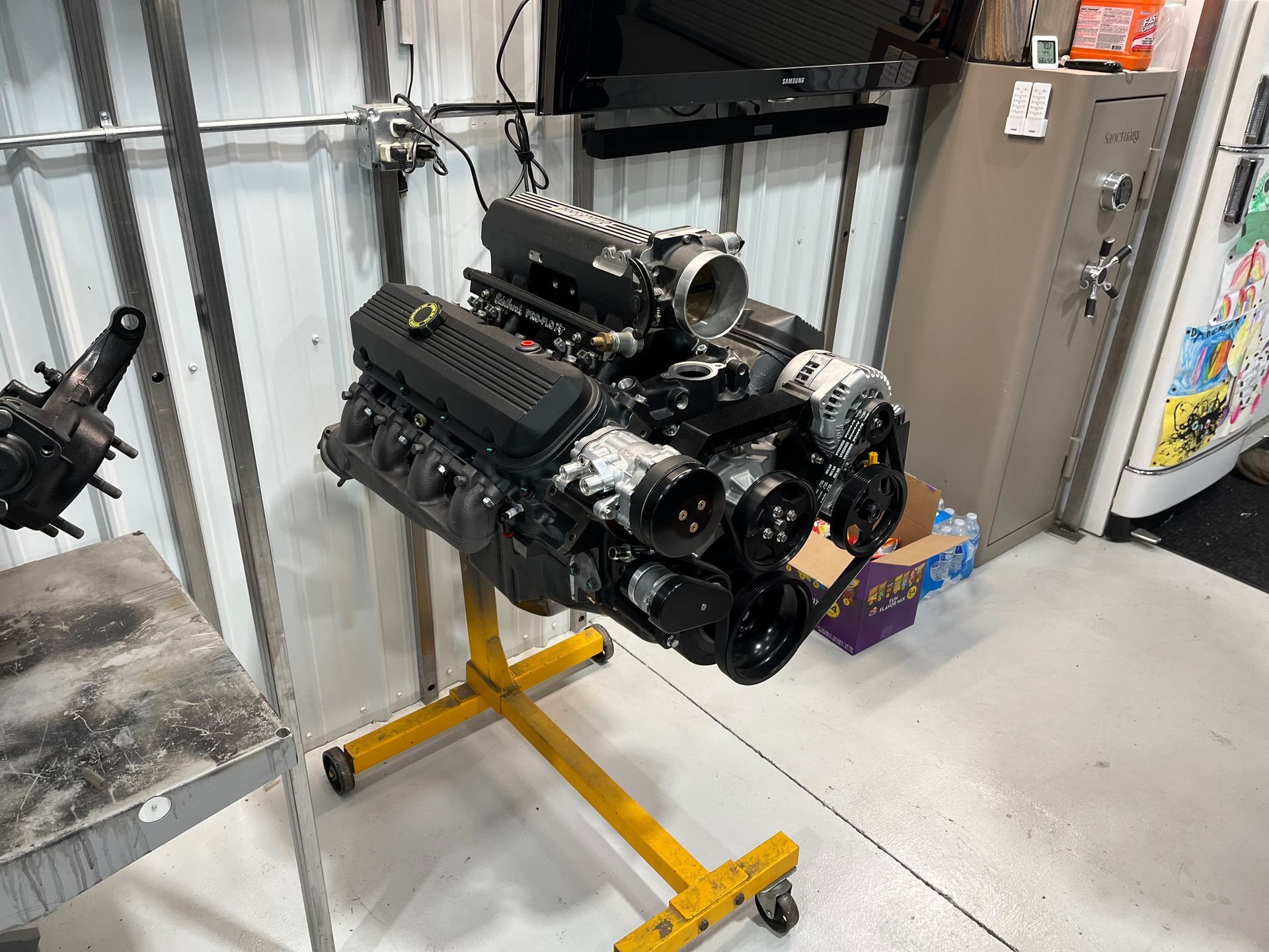 Engine on a yellow stand in a workshop garage, with corrugated metal walls and a refrigerator nearby