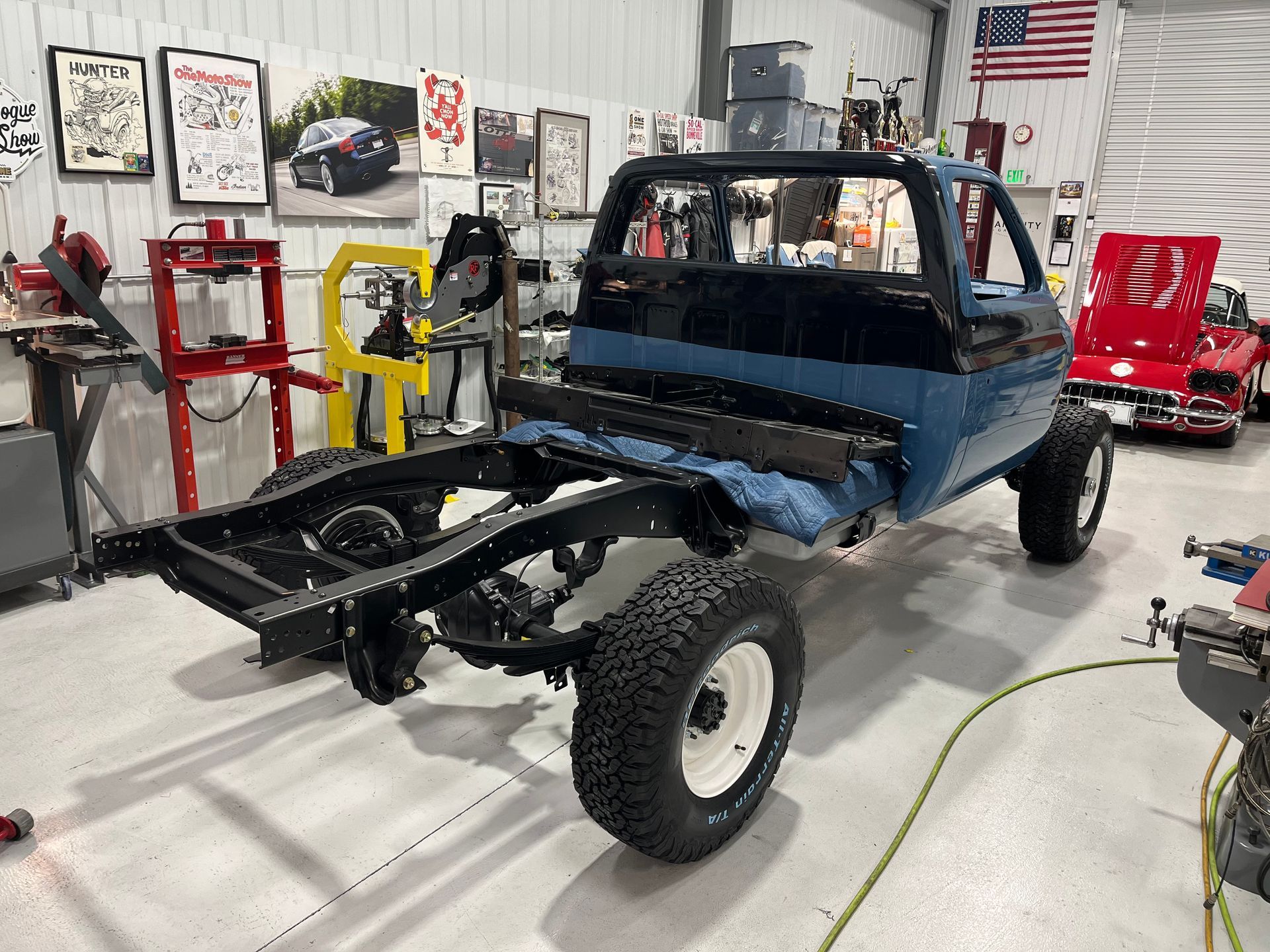 Partially restored blue pickup truck chassis in a workshop with tools and wall posters