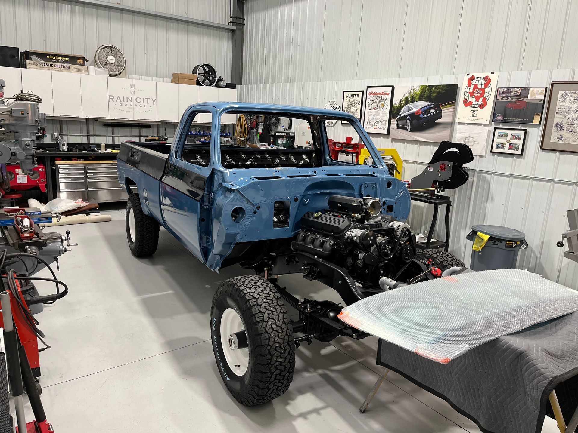 Blue pickup truck chassis in a garage, with exposed engine and off-road tires under repair