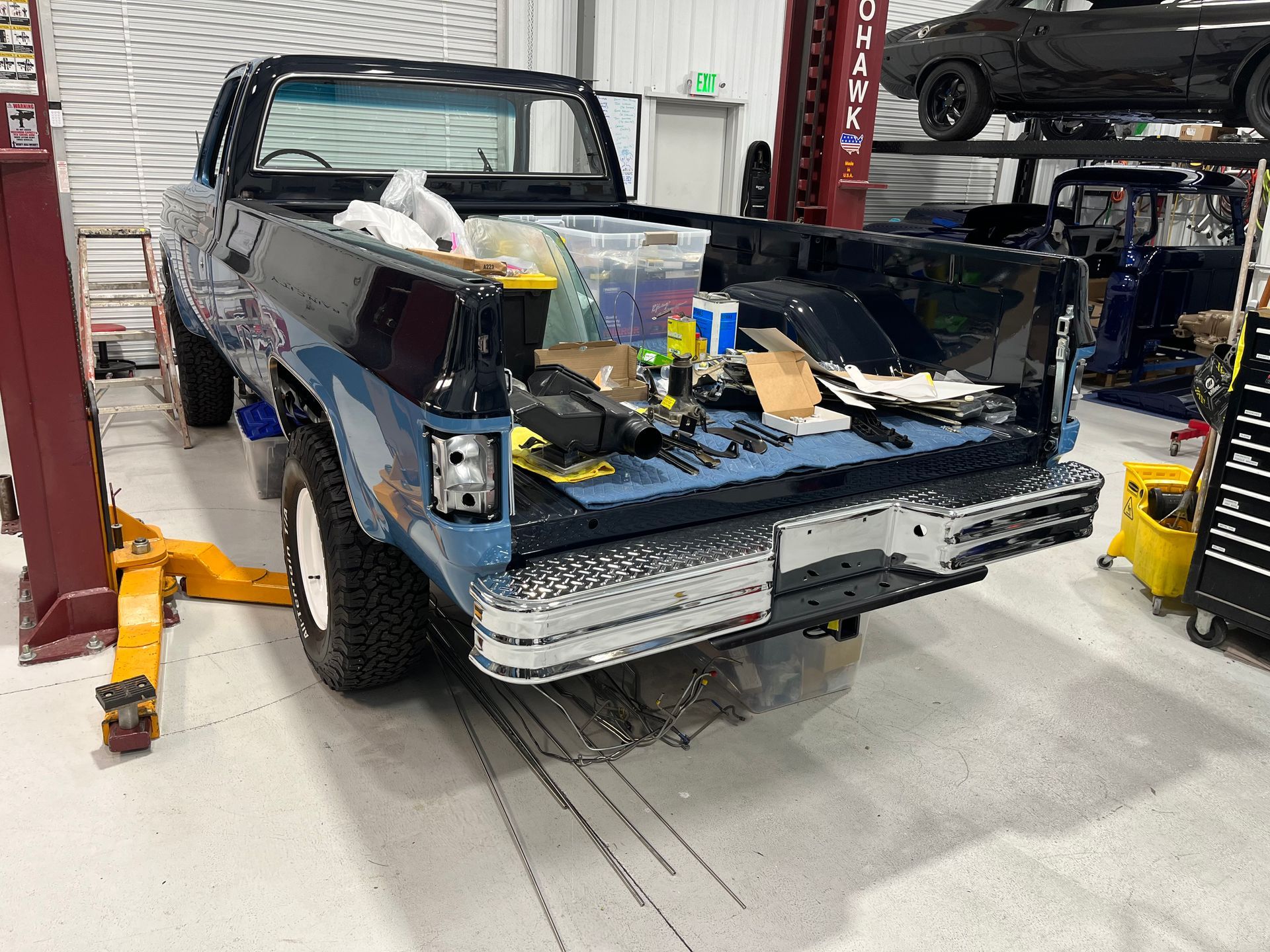 Partially disassembled dark pickup truck in an auto repair shop, rear bed exposed, tools nearby