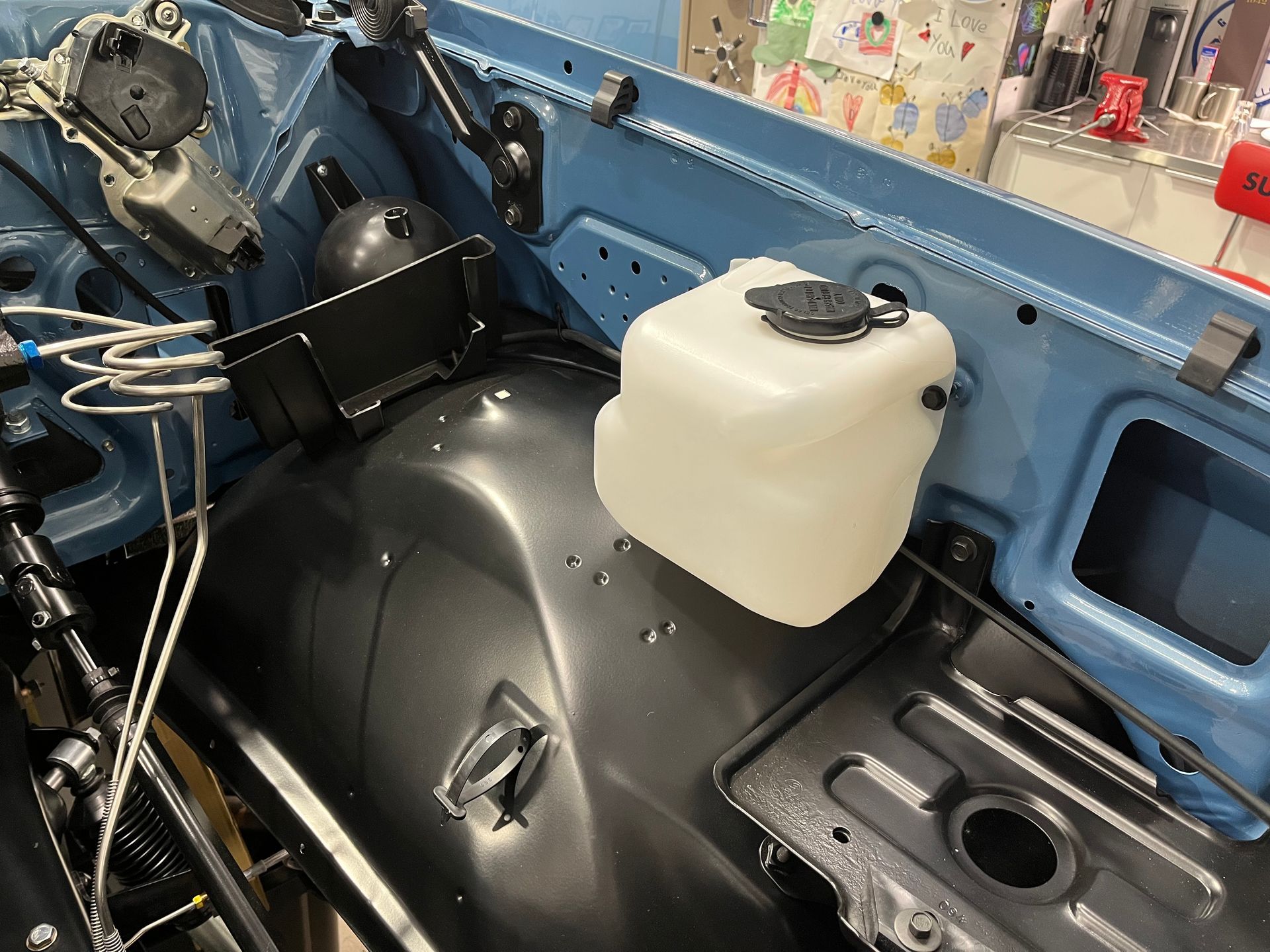 Car engine bay with a white coolant reservoir and black components inside a blue body shell