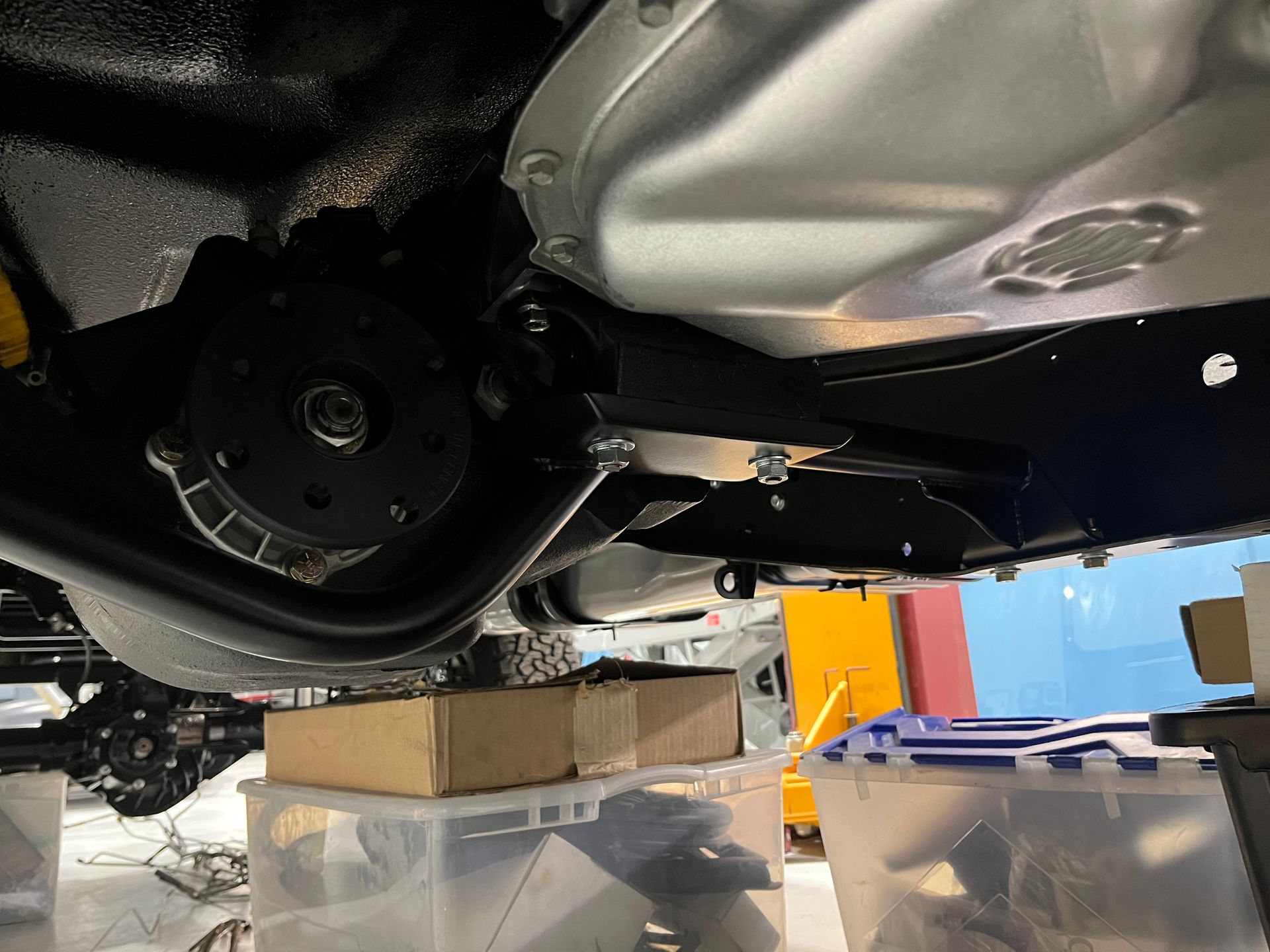 Underside of a vehicle on a lift, showing the rear axle and nearby boxes in a garage-like setting