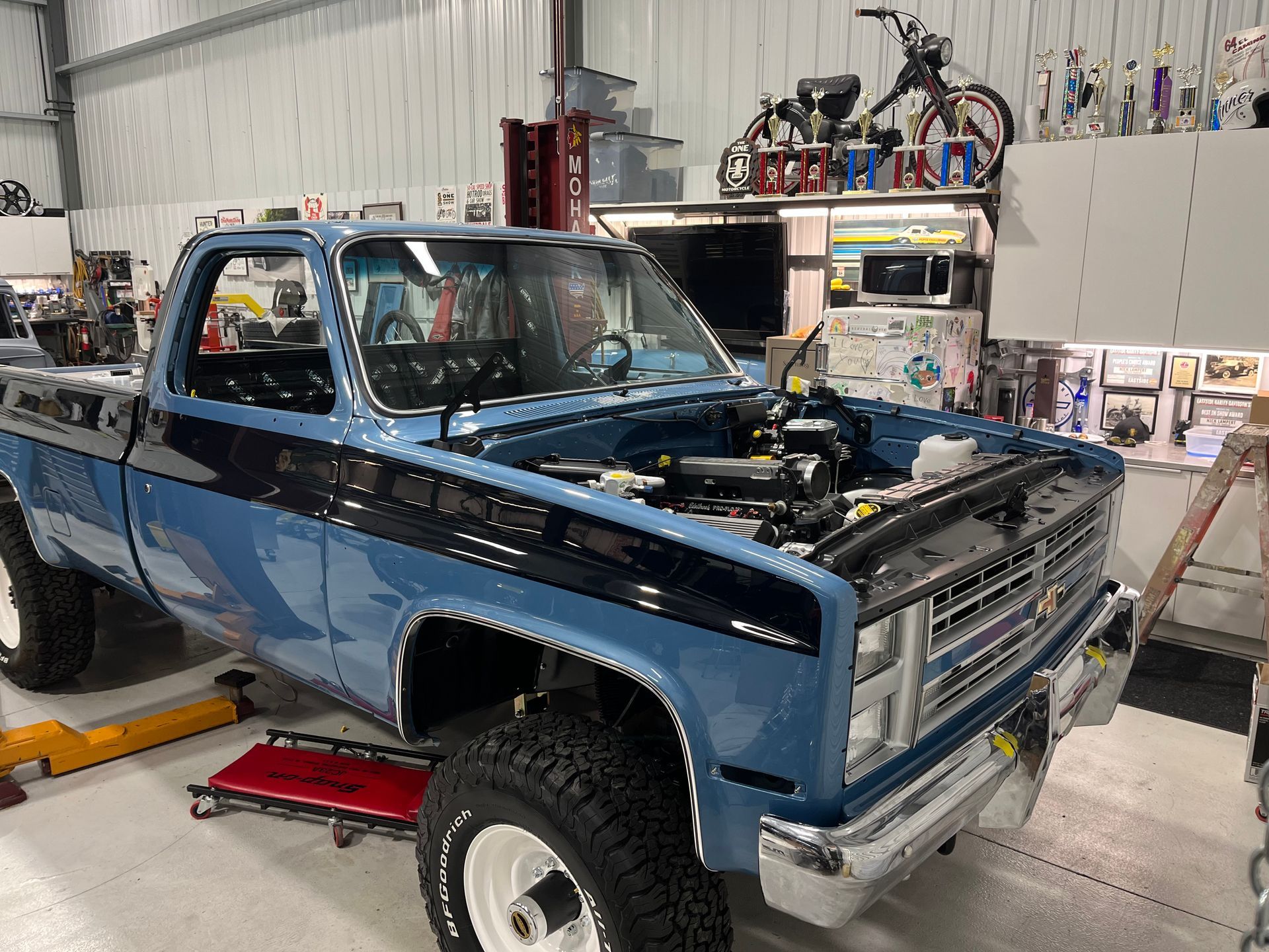 Blue pickup truck with missing hood in a workshop garage, surrounded by tools and shelves