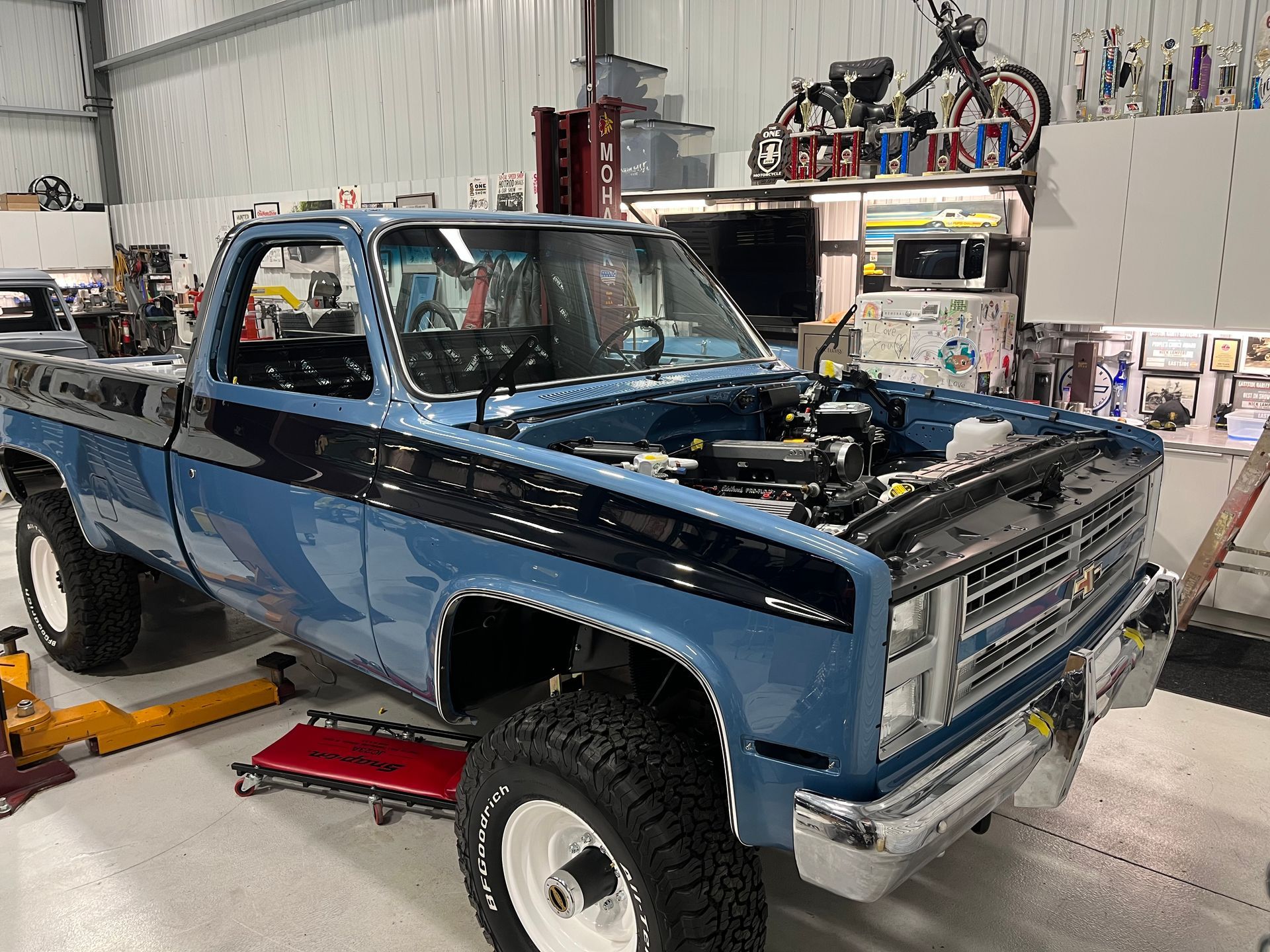 Blue pickup truck under repair in a workshop, front hood open and lifted on jacks.