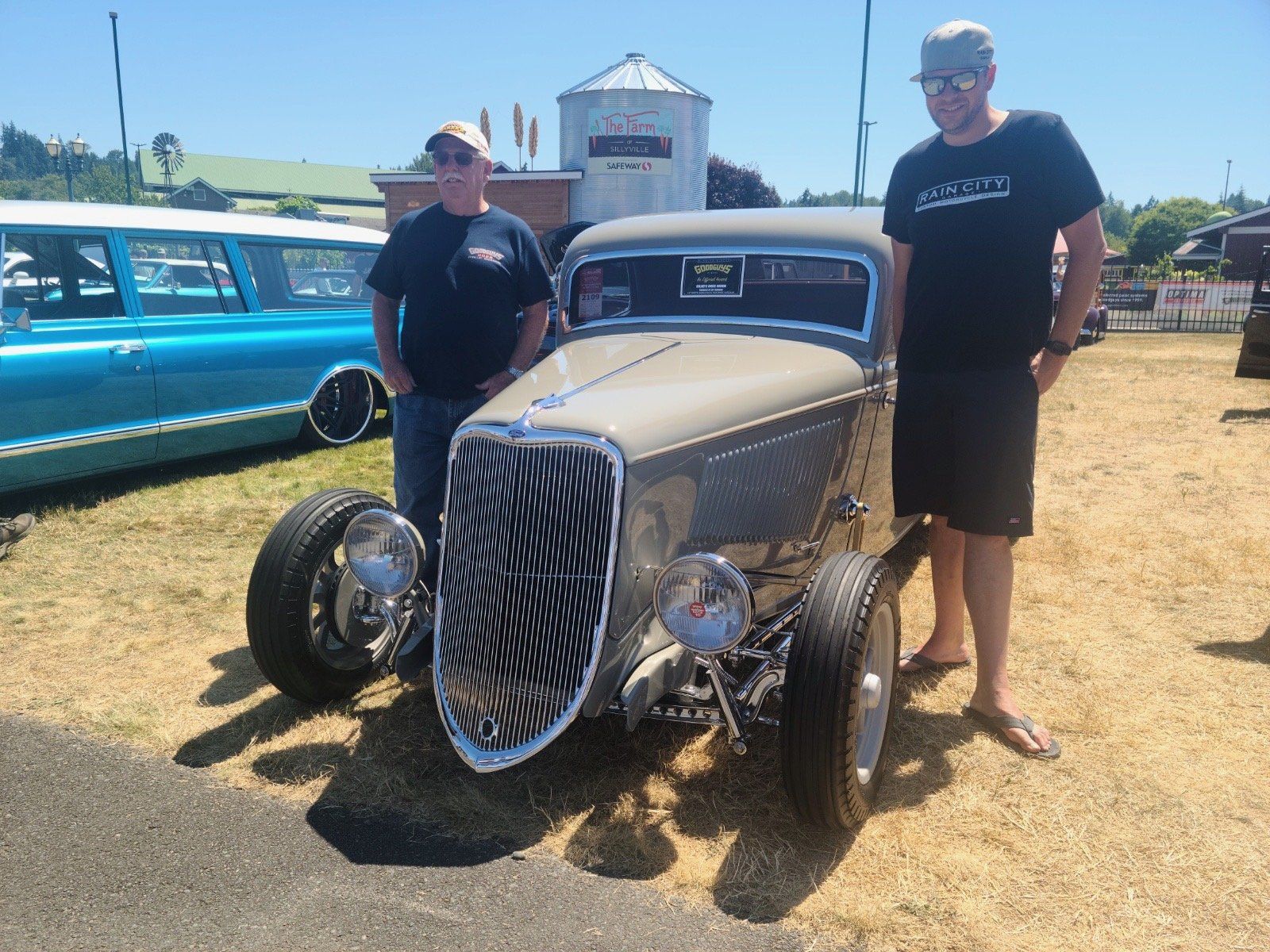 Two men standing beside a tan vintage hot rod at an outdoor car show