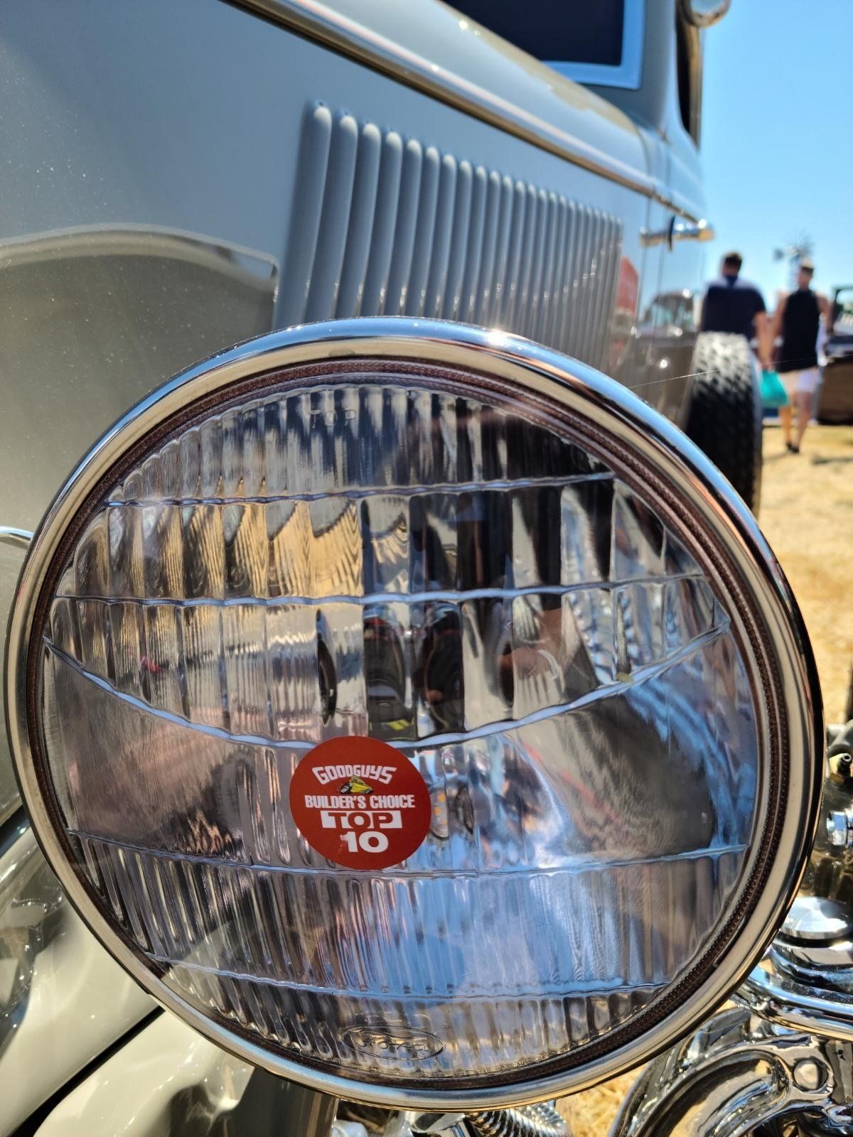 Close-up of a chrome vintage car headlight with a red sticker, parked outdoors on a sunny day