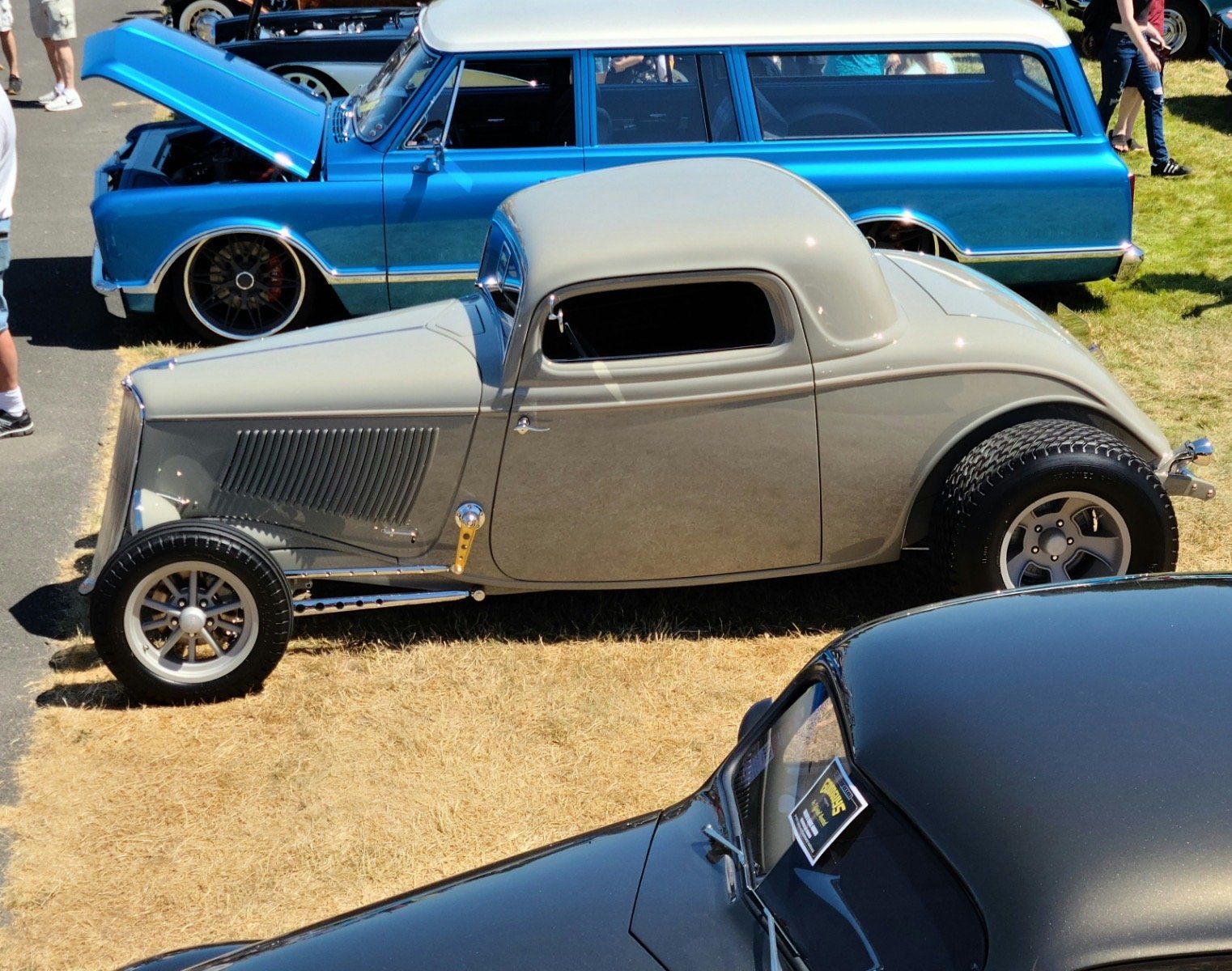 Classic gray hot rod car at an outdoor car show, with a blue station wagon and other vehicles nearby