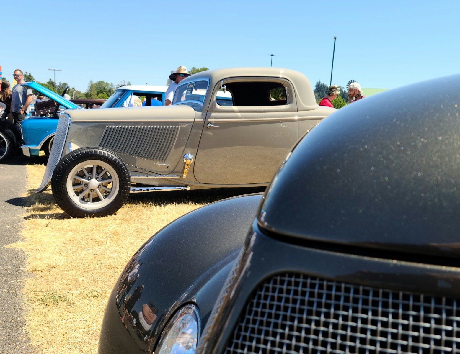 Classic cars at an outdoor car show, including a tan hot rod and a black car in the foreground.