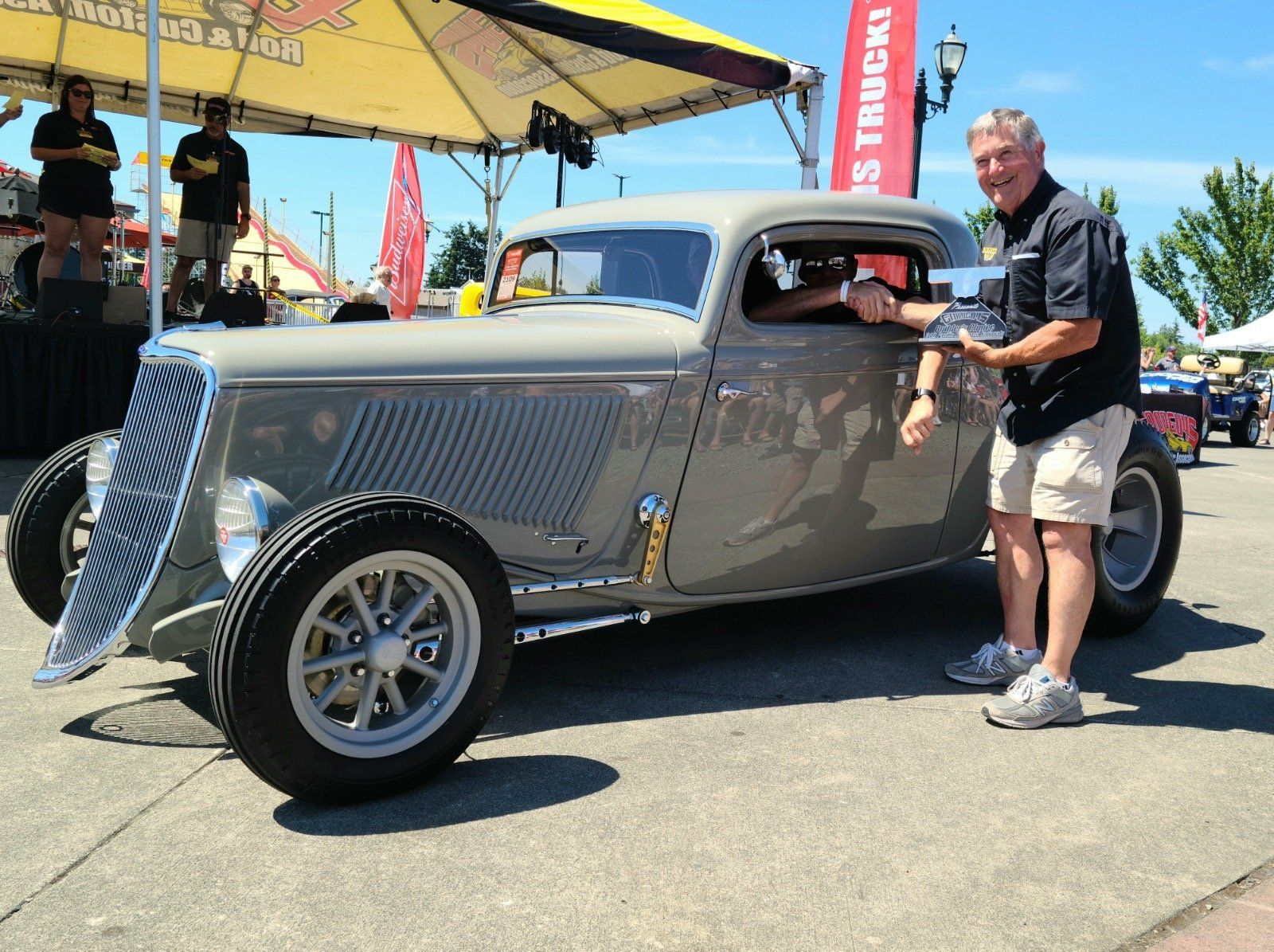 Gray classic hot rod car at an outdoor car show, with a man standing beside it under a yellow canopy.
