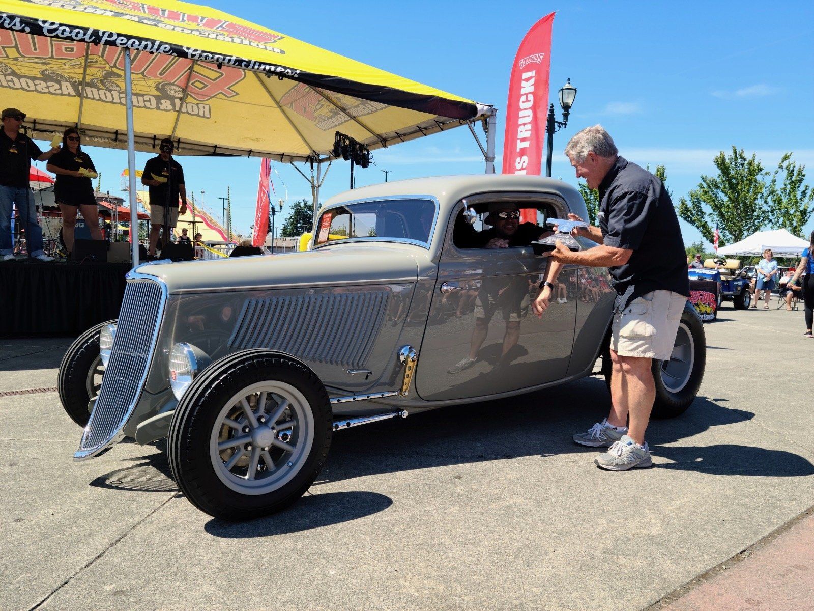 Classic silver hot rod at outdoor car show, man leaning into the driver’s window.