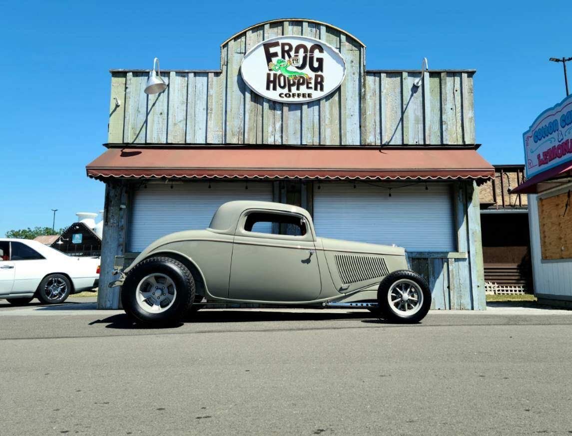 Vintage hot rod parked in front of a weathered Frog Hopper storefront under a clear blue sky