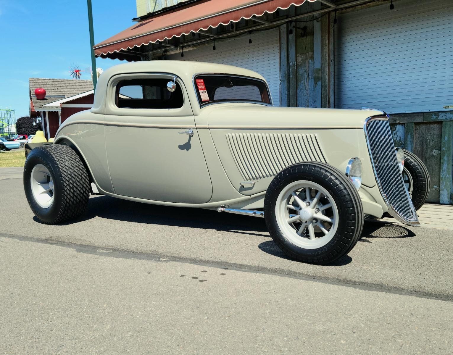 Vintage beige hot rod parked on a sunny street beside a blue building.