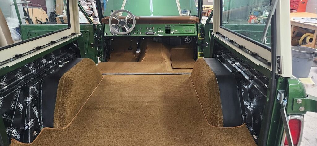 Interior of a green vintage car with brown carpet and black side panels, viewed from the back seat area