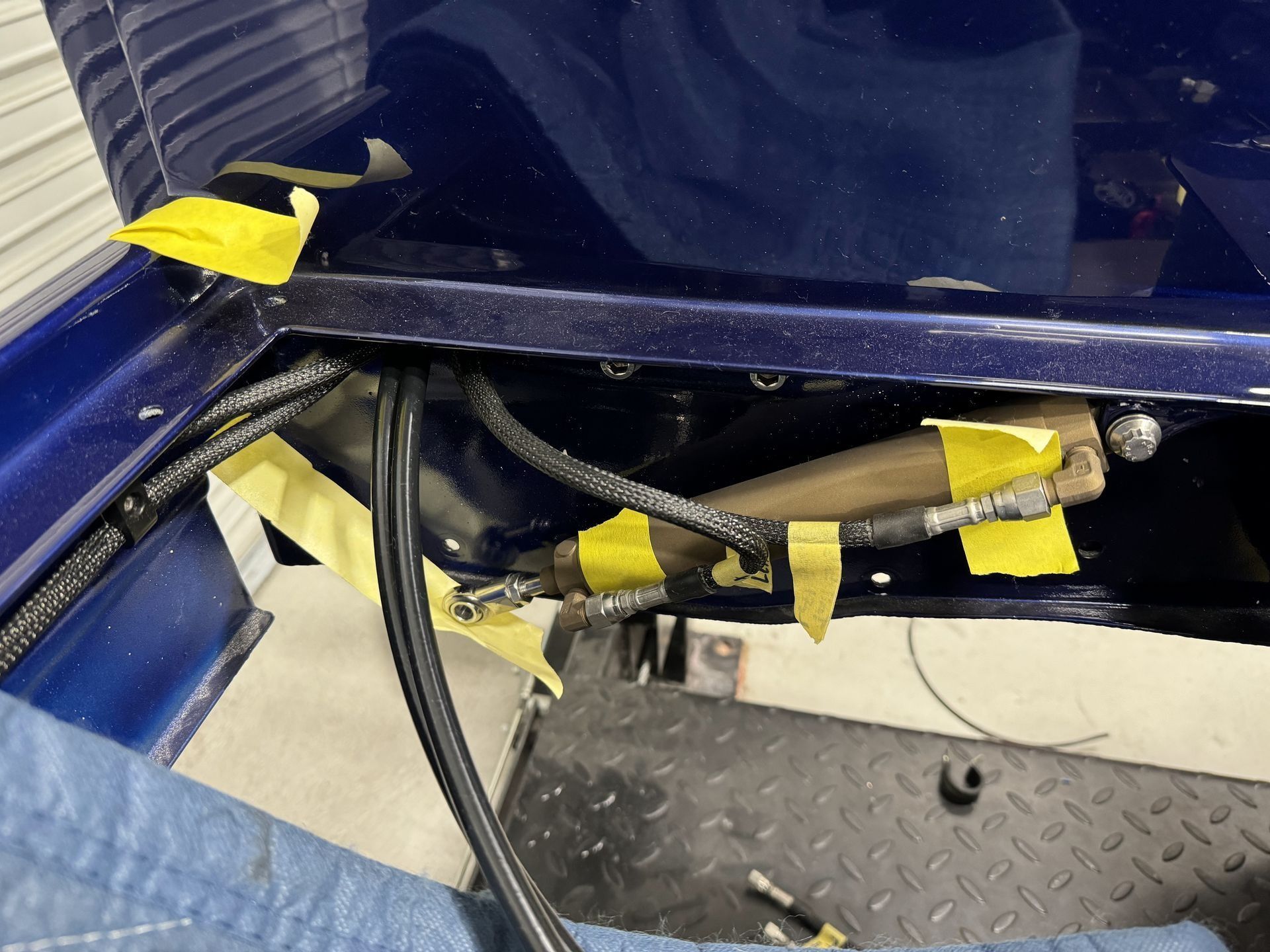 Close-up of exposed wiring and taped components under a blue vehicle panel, with a diamond-plate floor below