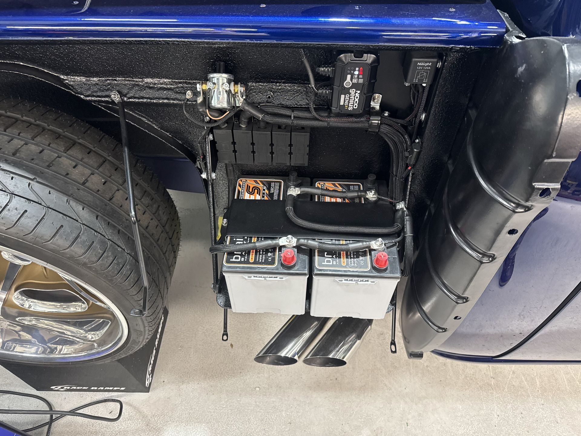 Battery compartment mounted beneath a blue vehicle, with exposed wiring and components near the rear wheel
