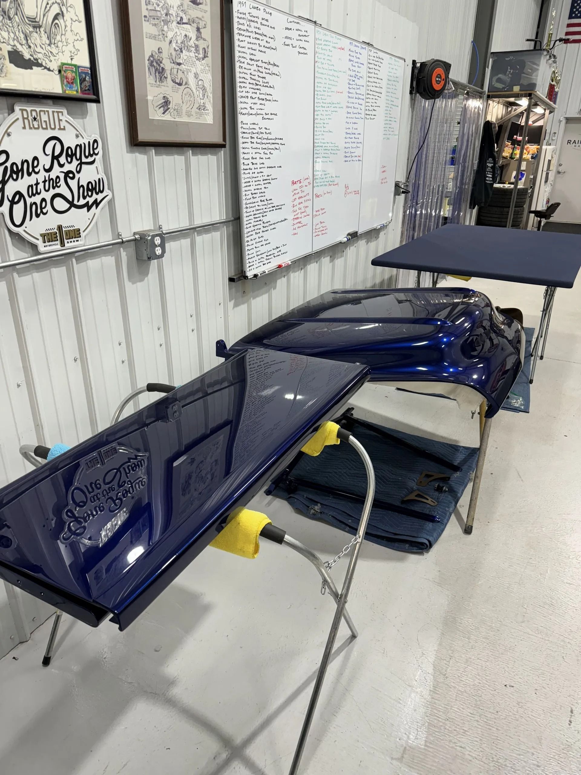 Blue car body panels stacked on a workbench in a garage or workshop with whiteboard notes nearby