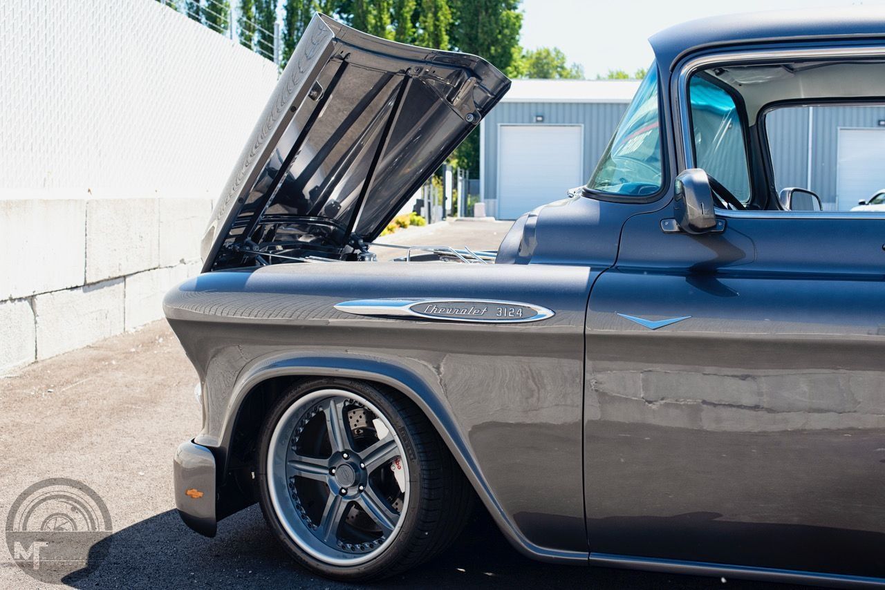 Classic dark blue pickup truck with hood open, parked beside a white wall in a sunny lot.