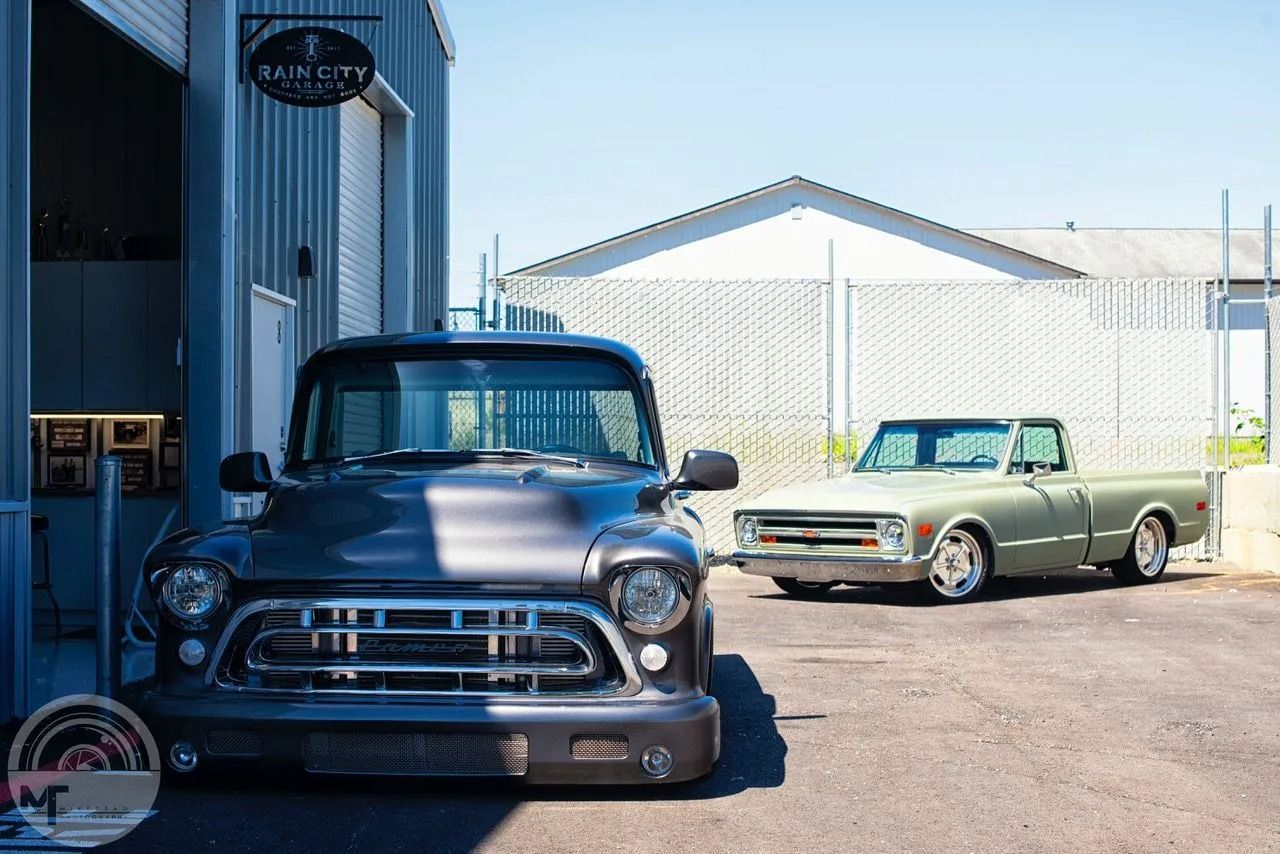 Two vintage pickup trucks parked outside a warehouse in bright sunlight.