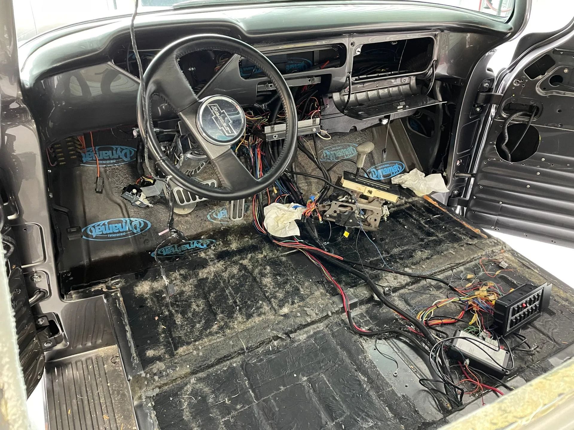 Inside a stripped car interior with exposed wiring, bare floor, and a steering wheel.
