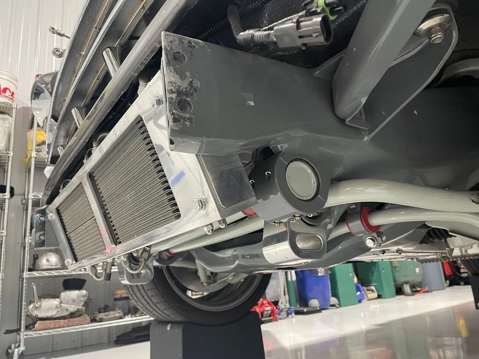 Underside of a vehicle in a repair shop, showing the exhaust, suspension, and a radiator-like component.
