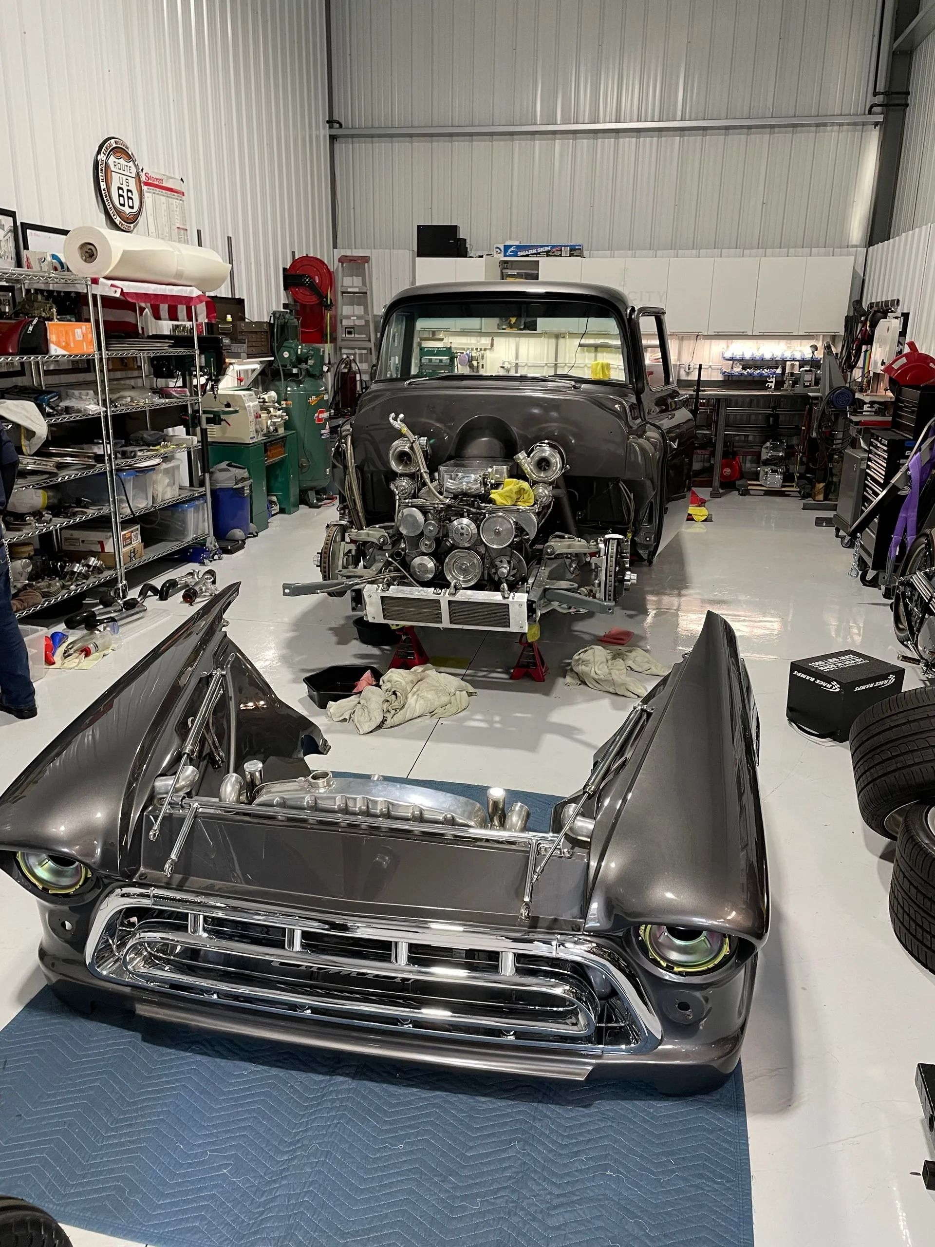 Classic car restoration shop with a partially disassembled black car and detached front end in the foreground.