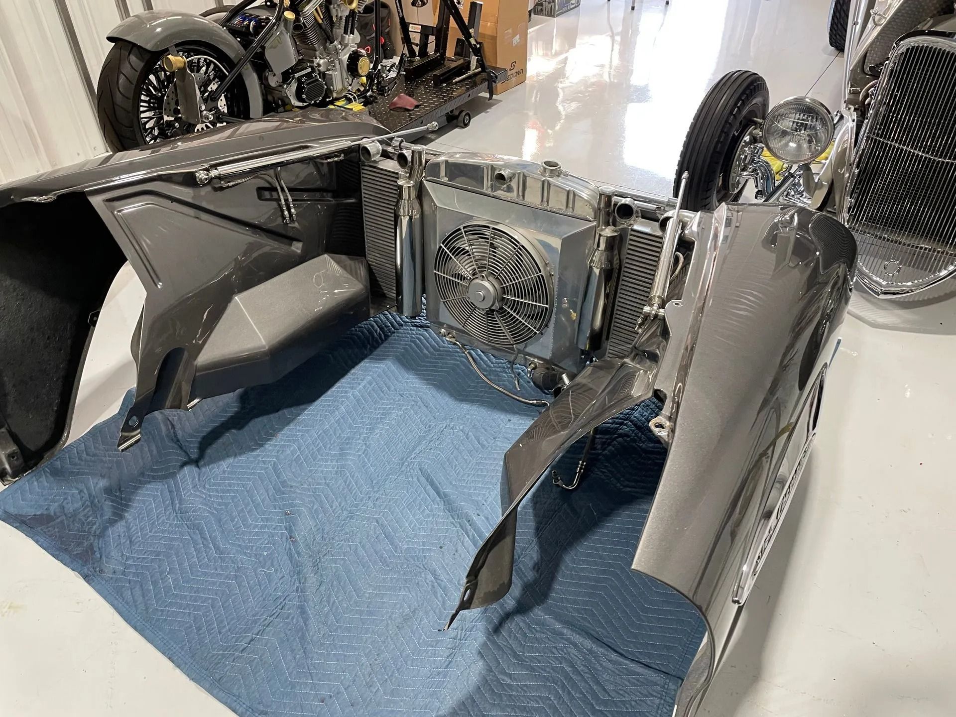 Silver race car body shell with exposed rear cooling fan and blue carpeted floor indoors