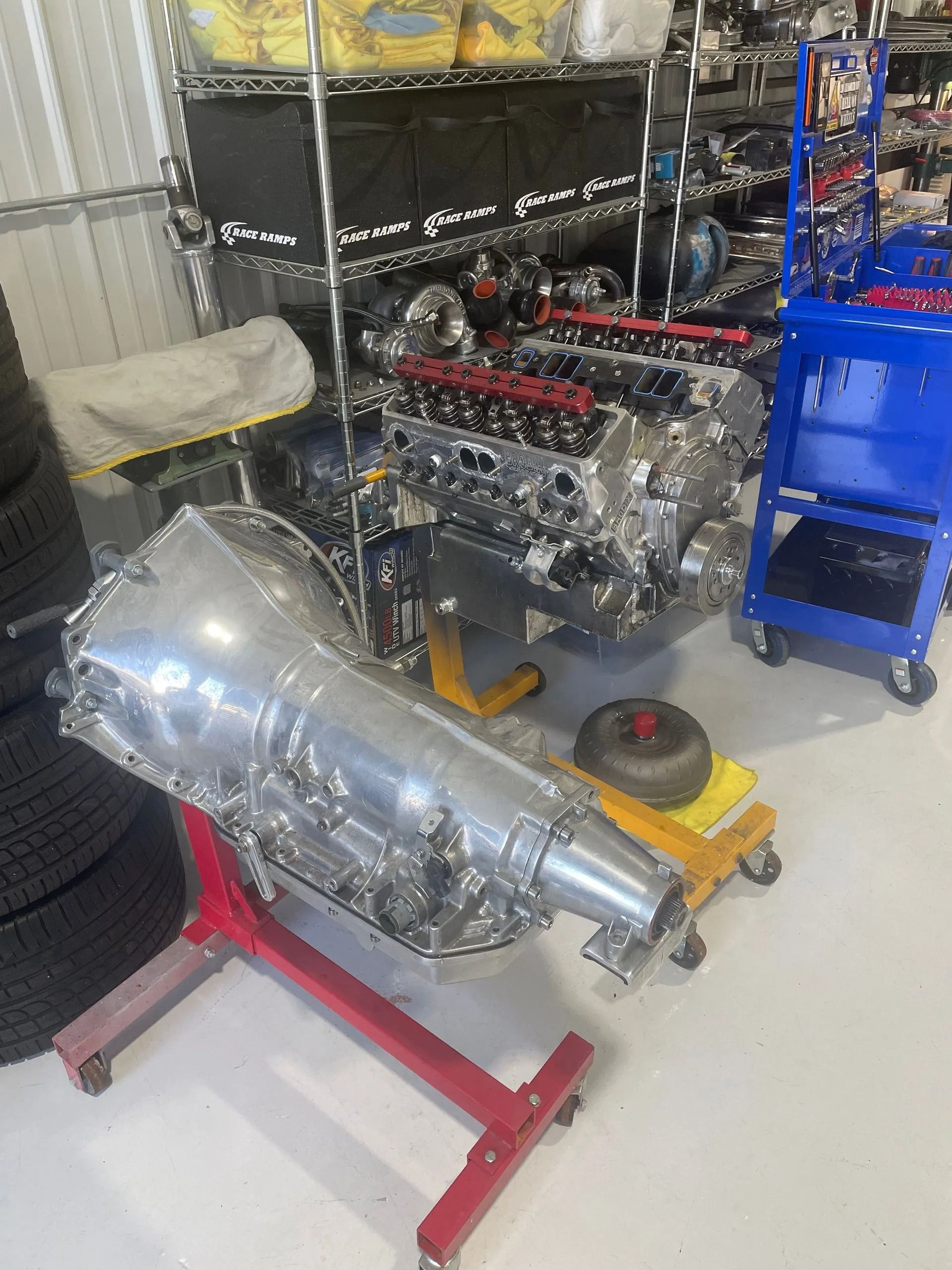 Classic car engine and transmission on shop stands in a garage, with tools and a blue cart nearby.