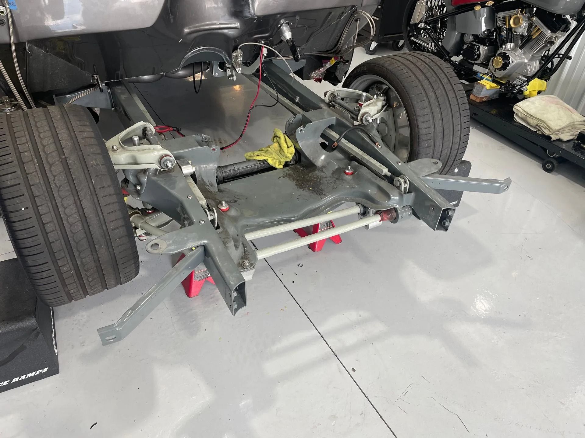 Car chassis on a garage floor with exposed suspension, wheels, and red jack stands