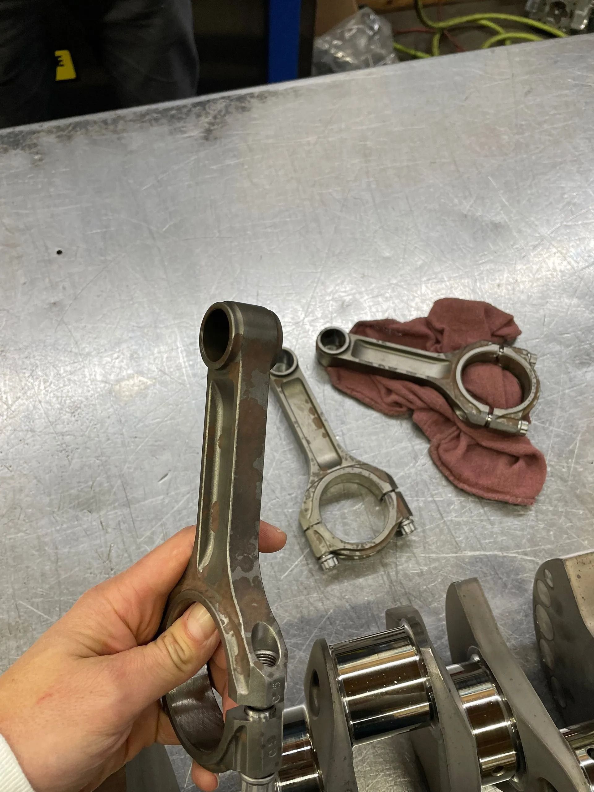 Hand holding a metal connecting rod beside an engine crankshaft on a workbench