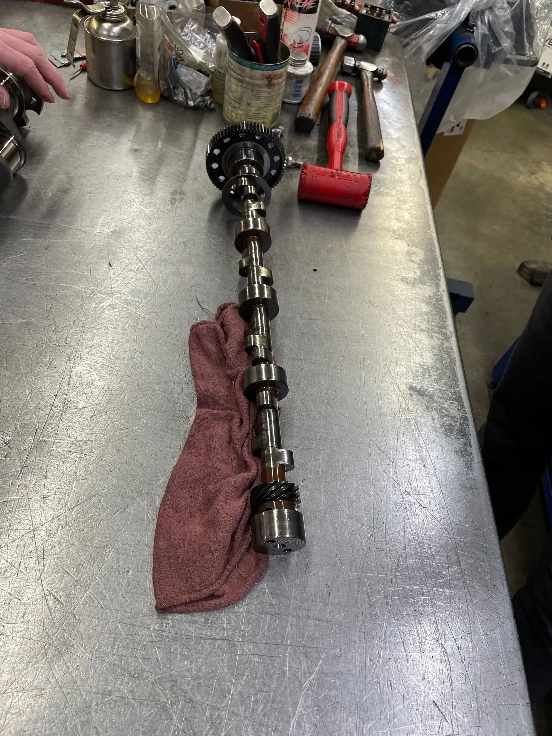 A long camshaft-like metal part on a workbench with a red towel beside it.