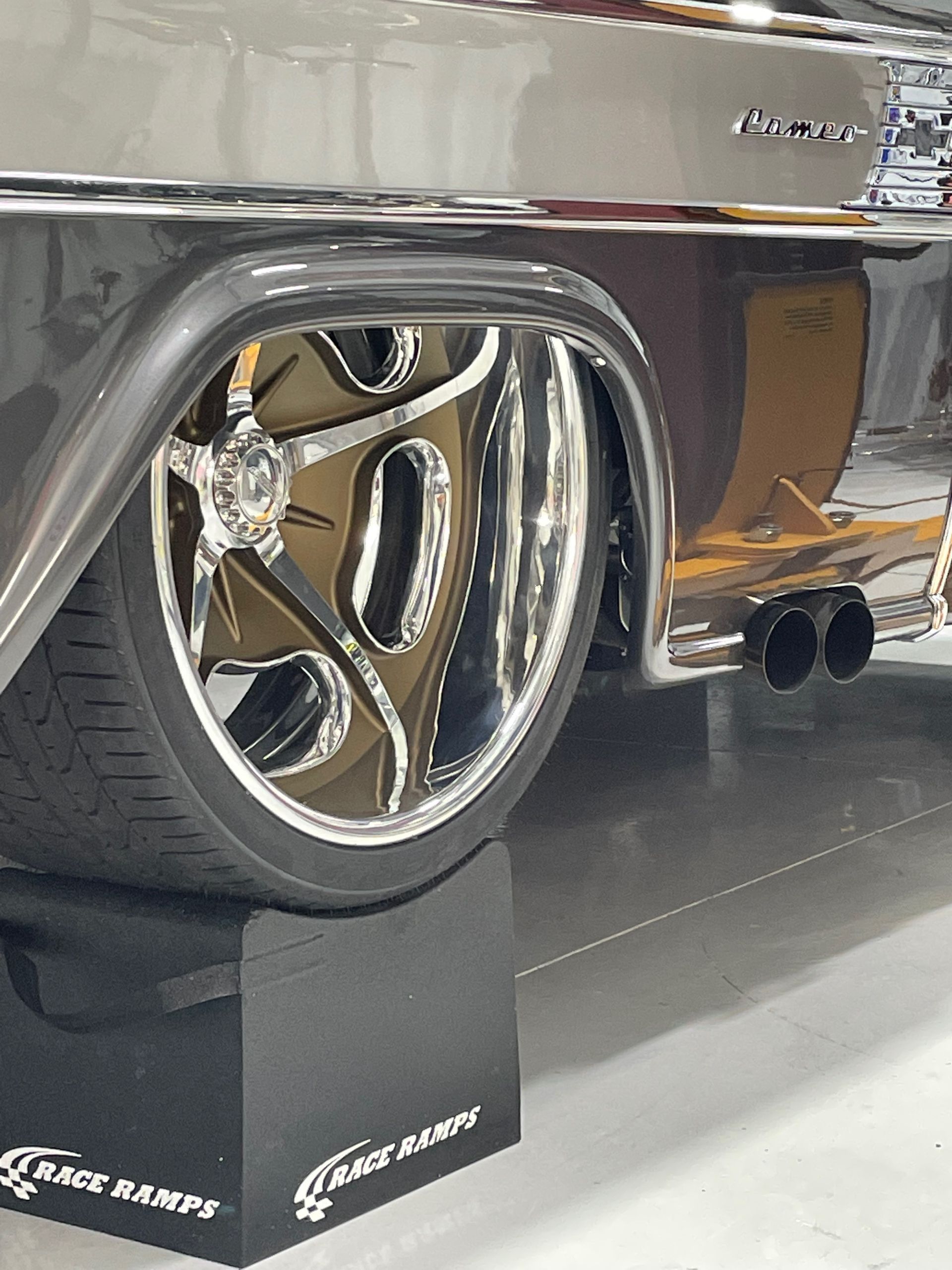 Polished classic car wheel and chrome body detail on display stand, with dual exhaust pipes nearby