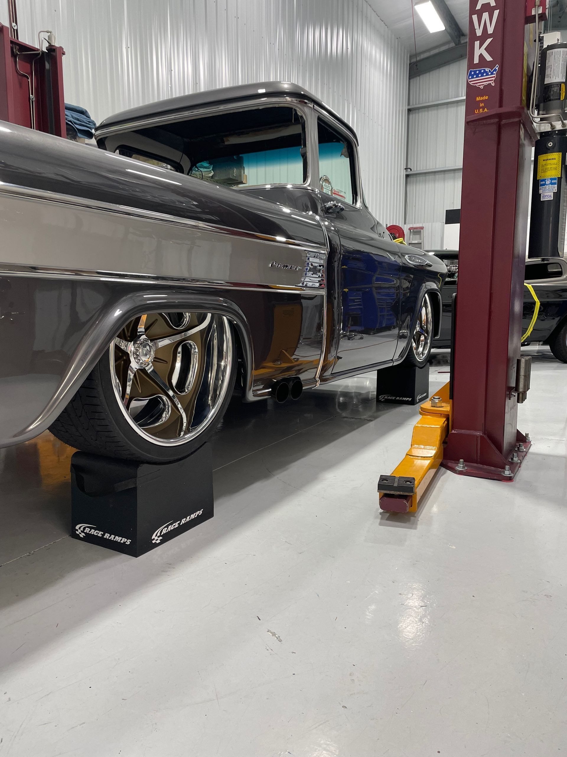 Classic gray-and-blue custom lowrider in a garage, raised on stands beside a red hydraulic lift.