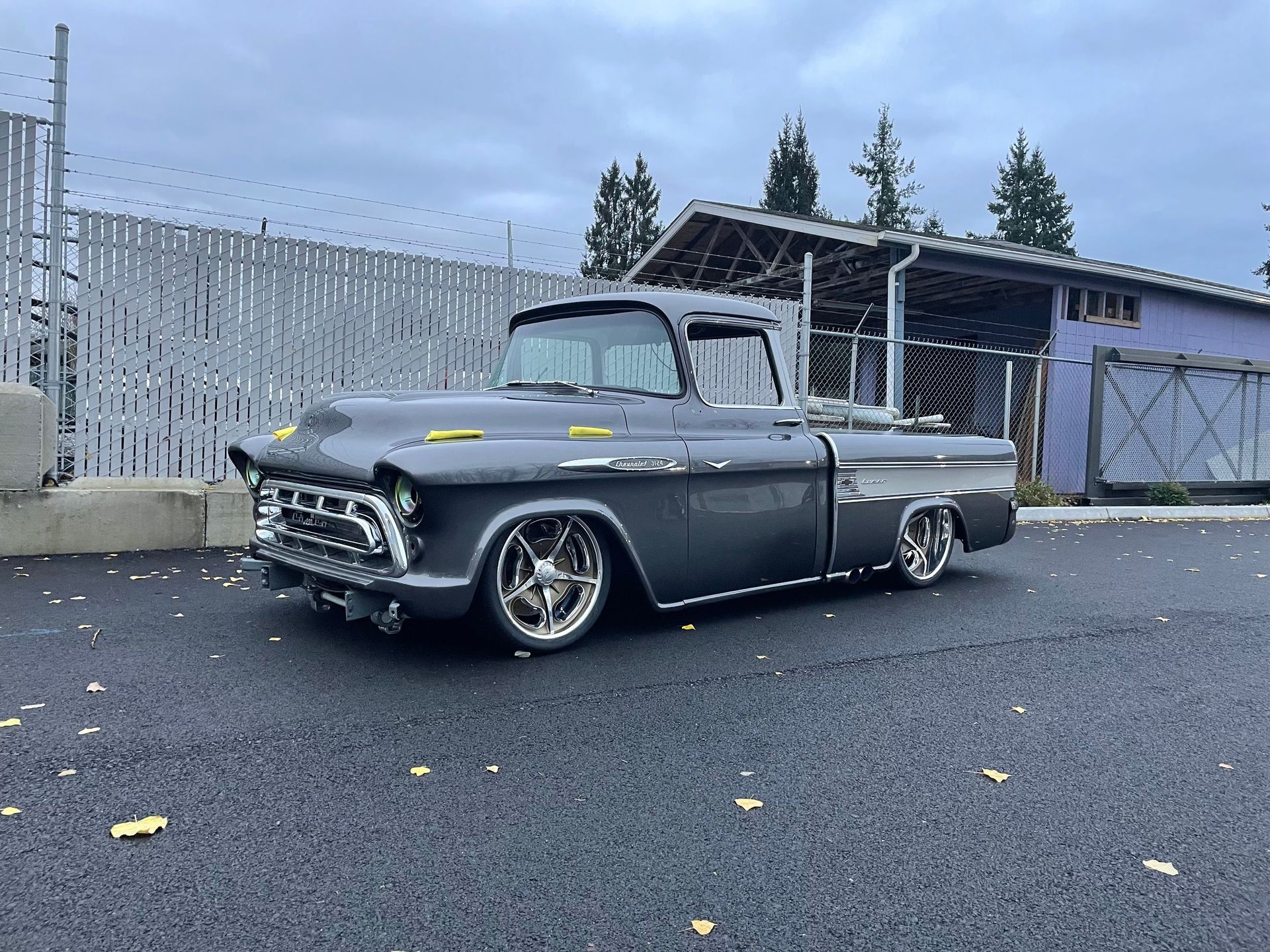 Classic gray lowrider car parked on wet pavement beside a fence and garage