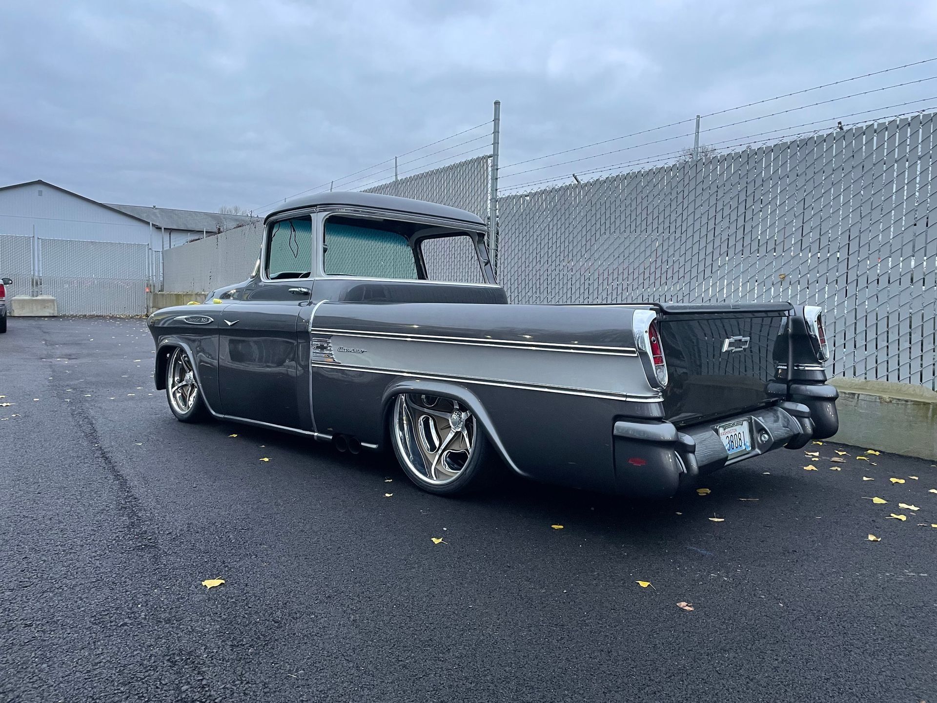 Classic black and gray lowrider pickup parked on a wet street beside a chain-link fence.