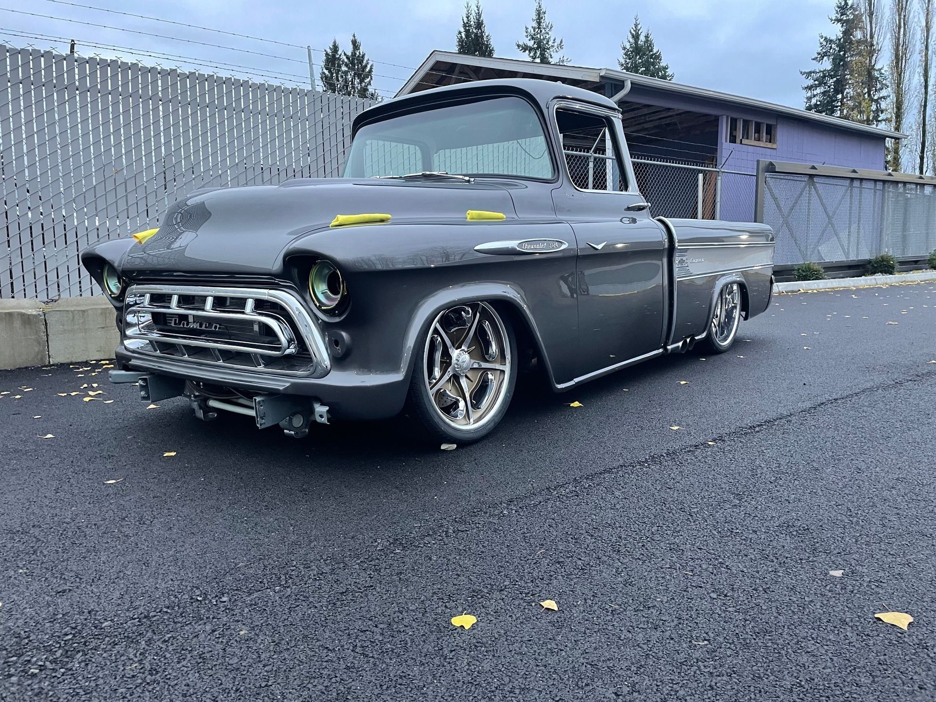Custom gray vintage pickup truck parked on a wet street beside a fence and building.