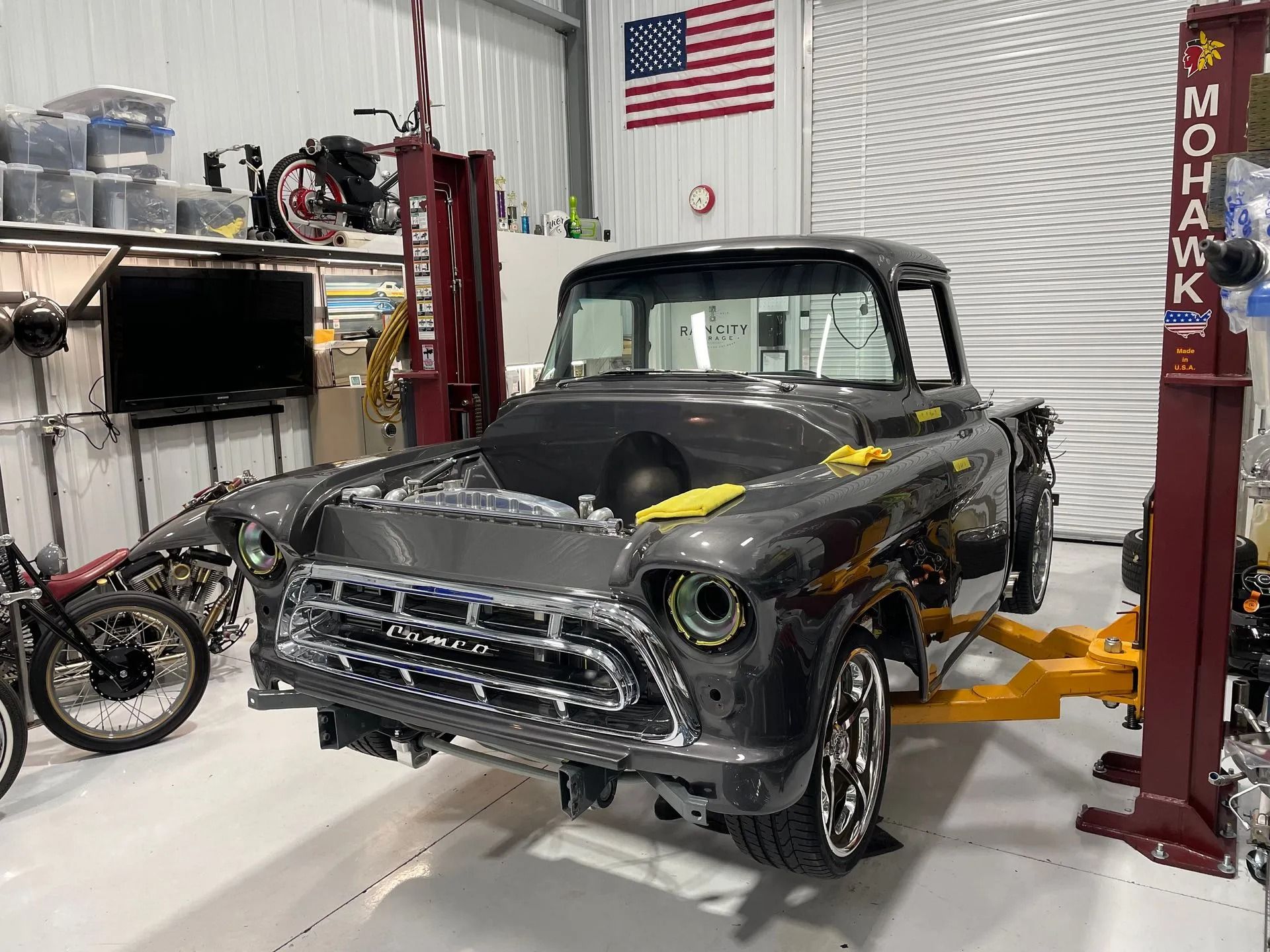 Classic black pickup truck in a garage on a lift, with a motorcycle and American flag in the background.