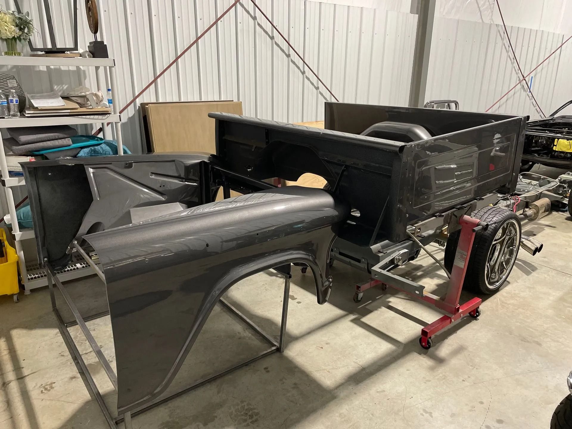 Black classic car body and parts in a workshop, with a front bumper on a stand and a vehicle frame on a jack.