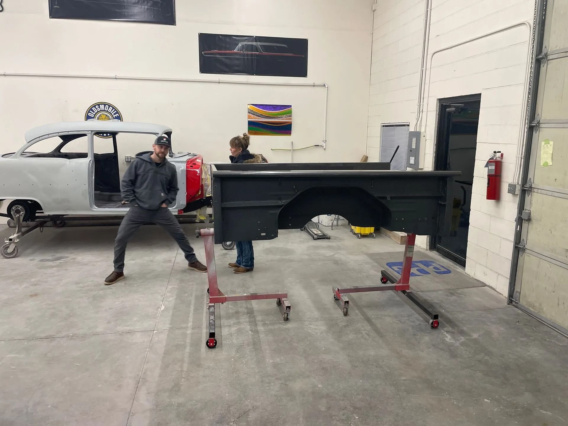 Two people in a workshop posing beside a large black metal frame on red stands.