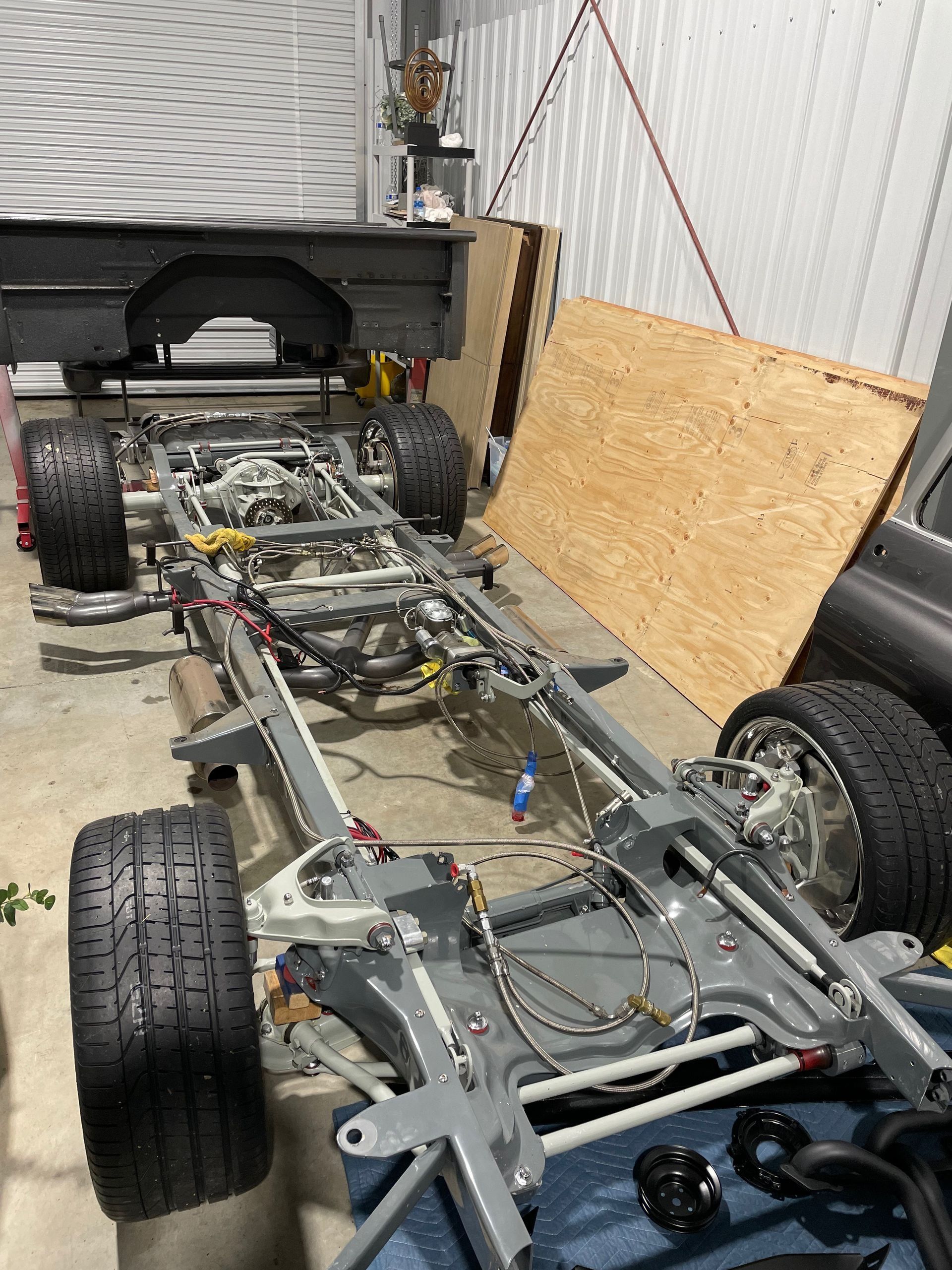 Partially assembled car chassis in a garage, with exposed frame, wheels, and plywood leaning nearby