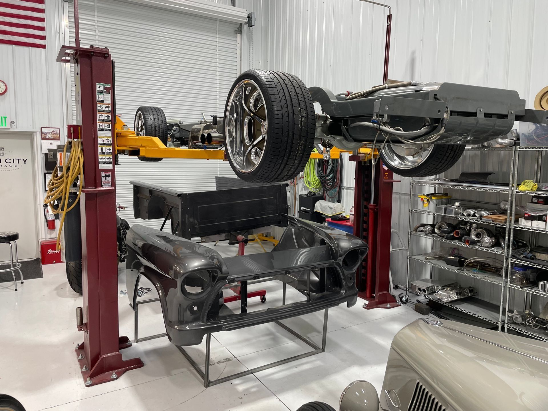 Auto shop with a car lifted on red hydraulic lifts, wheels removed, and parts arranged on shelves.