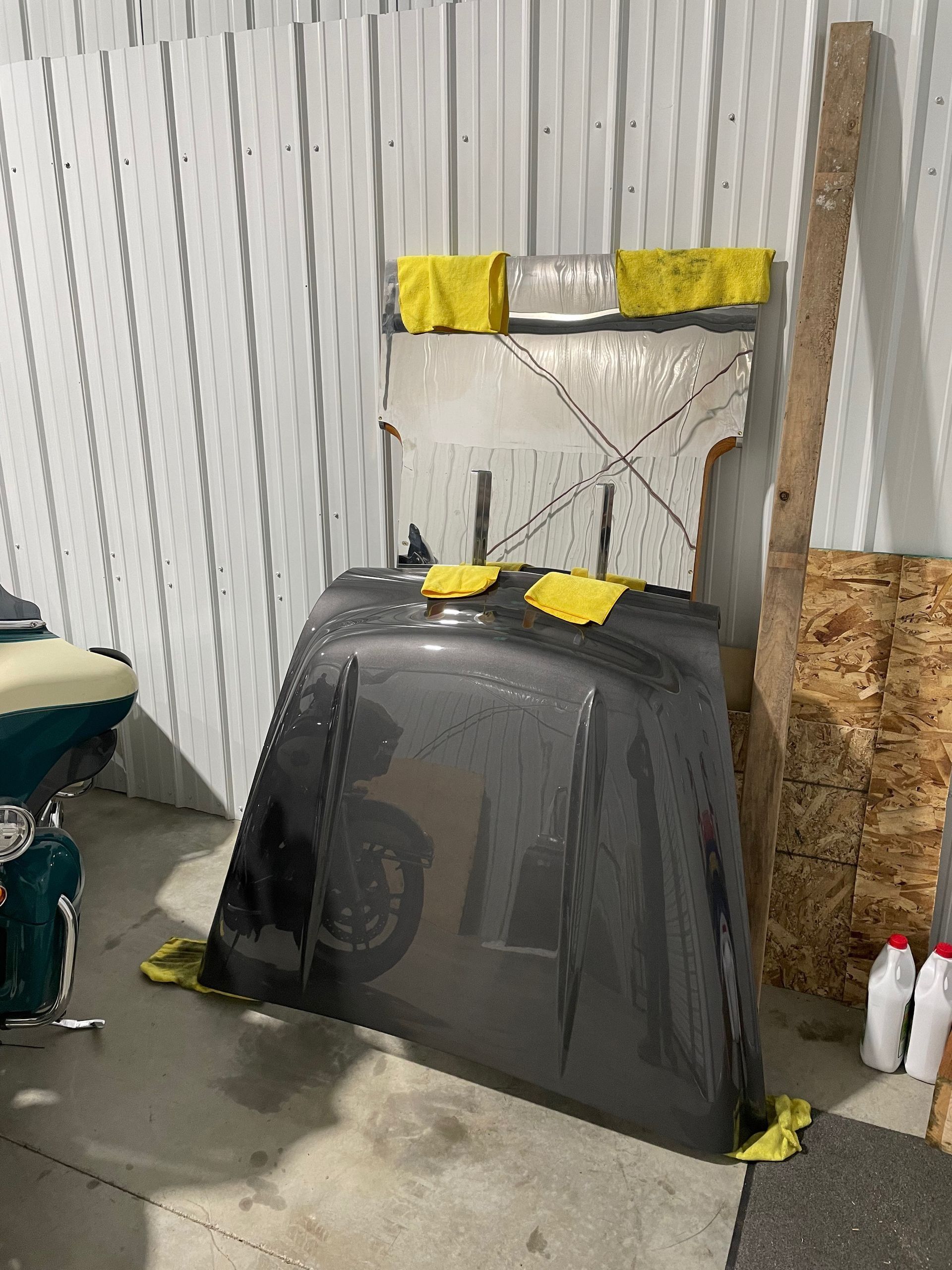 Black drying rack draped with translucent plastic, yellow tape, in a workshop beside a wicker chair and bottles