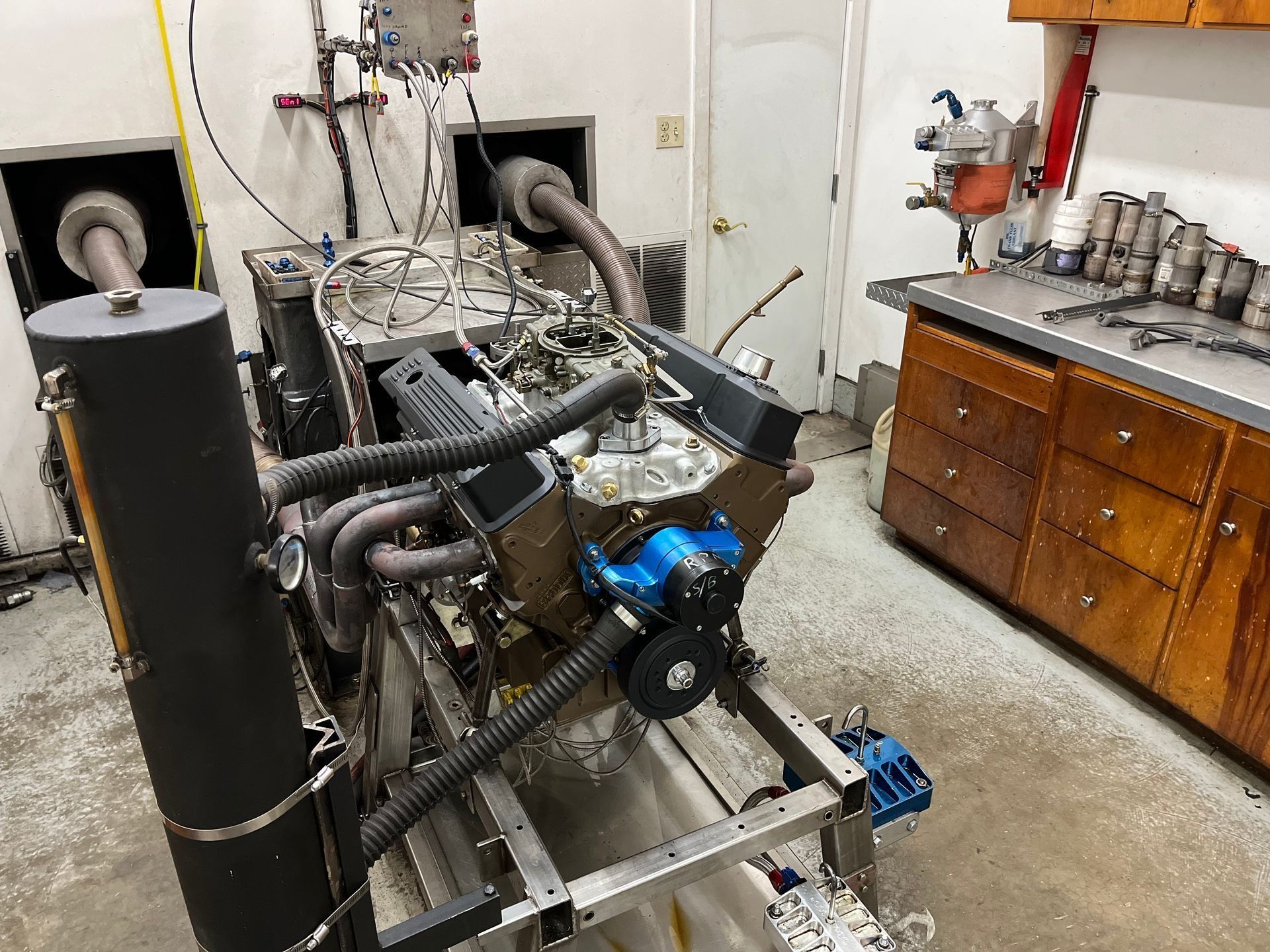 Industrial machinery on a metal frame in a workshop, with hoses, blue motor parts, and a wooden cabinet in the background