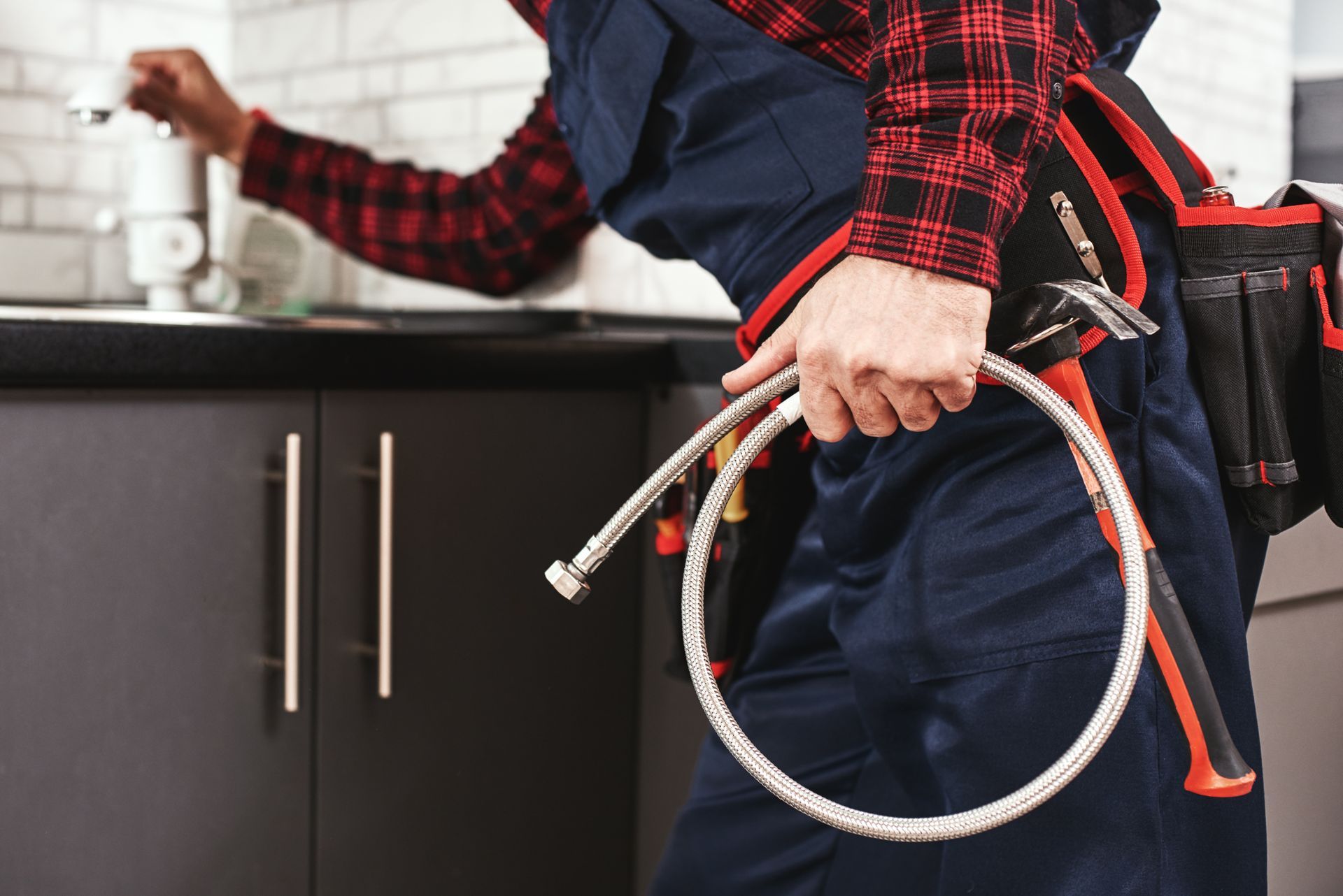 Plumber inspecting faucet, holding a flexible water supply line. Wearing work clothes, tool belt. Kitchen setting.