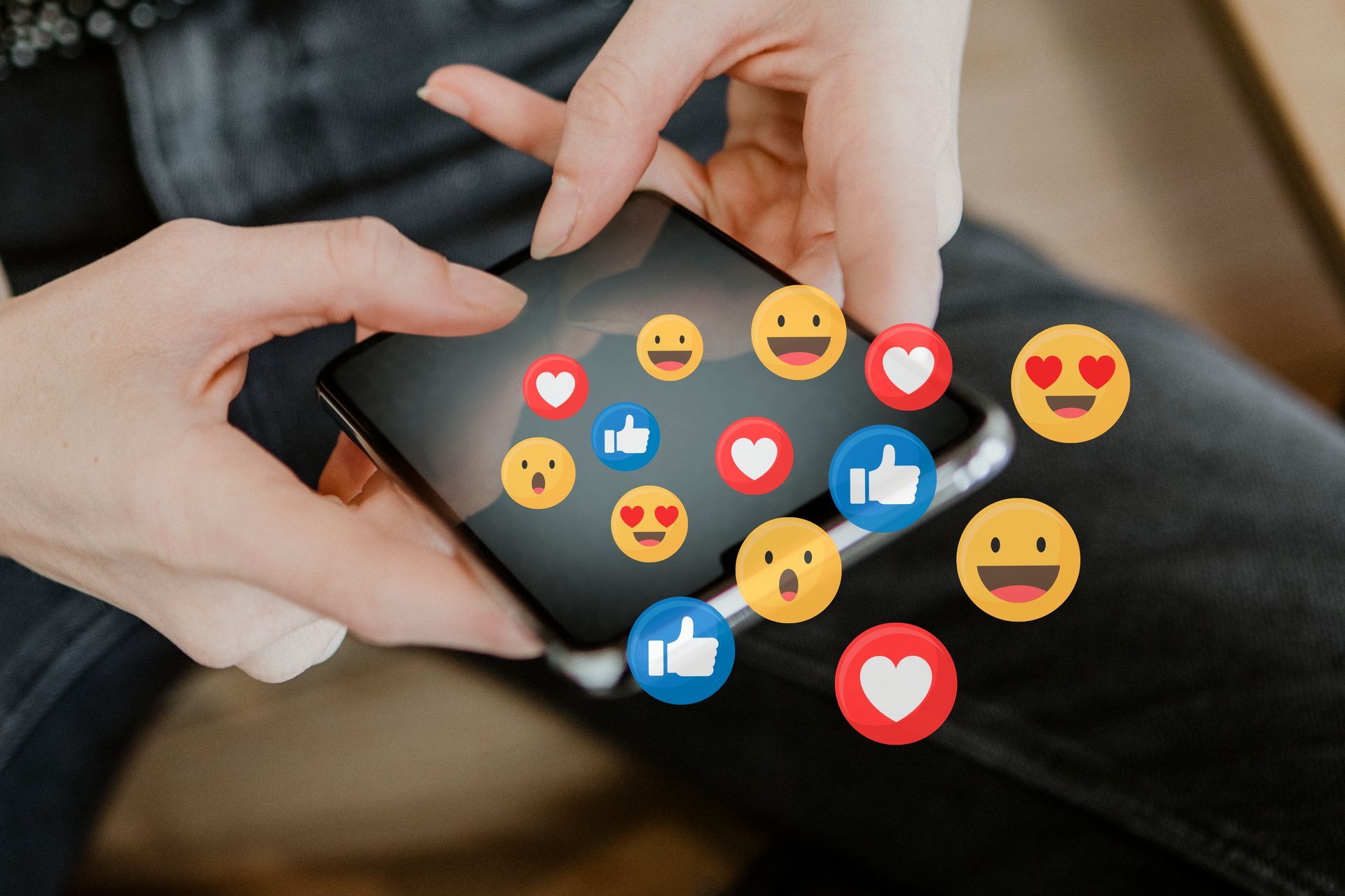 Hands holding a phone with animated emojis: hearts, thumbs up, and smiling faces.