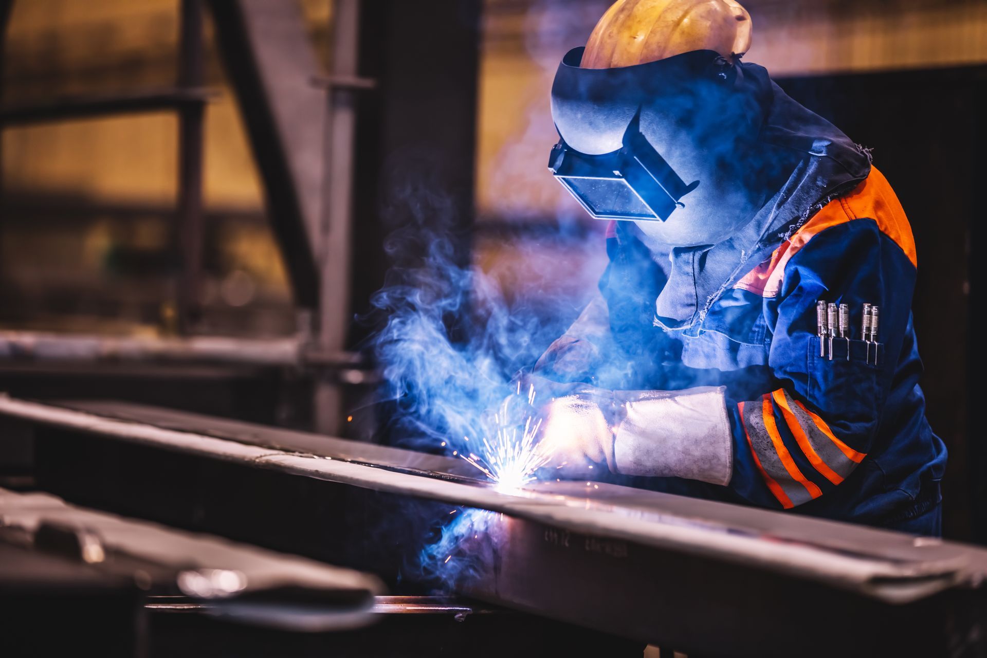 Welder in protective gear, welding metal beam with bright blue arc in workshop.