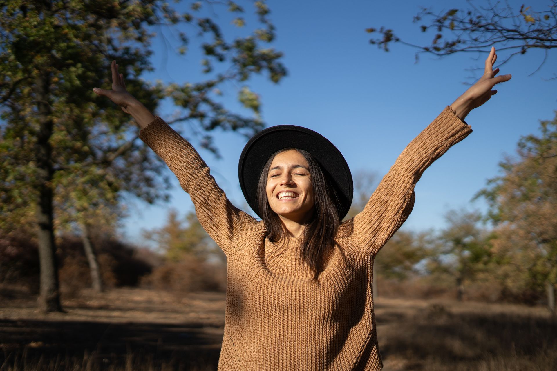 Woman wearing a hat with arms raised in joy, smiles outdoors with trees and blue sky.