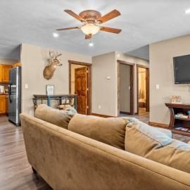 Living room with tan sectional sofa, ceiling fan, wall-mounted TV, and open doorway to hall and kitchen.